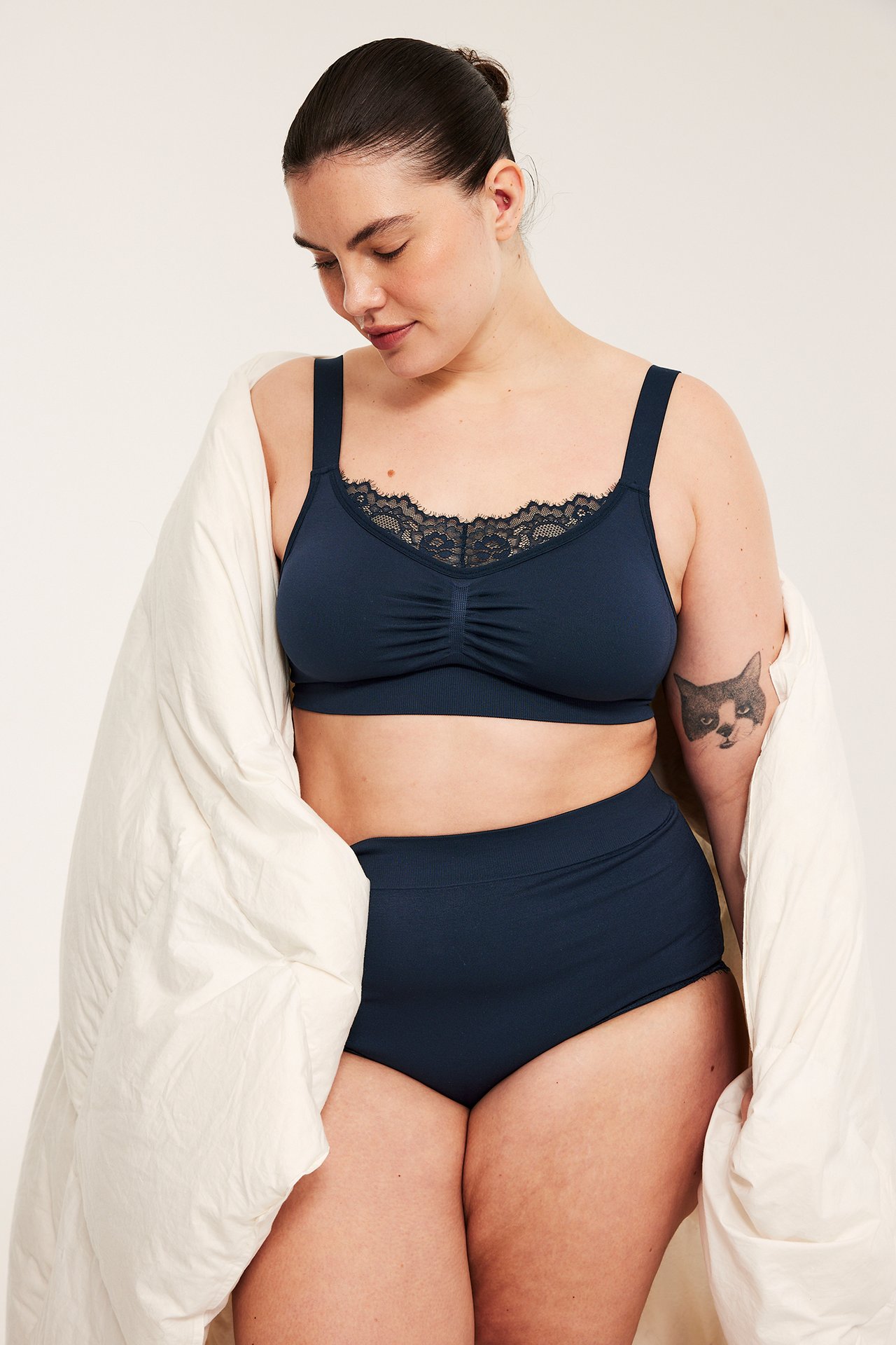 Frontvy av en kvinna i mörkblå sömlös bygellös bh med spets, matchande trosor och naturvitt täcke.