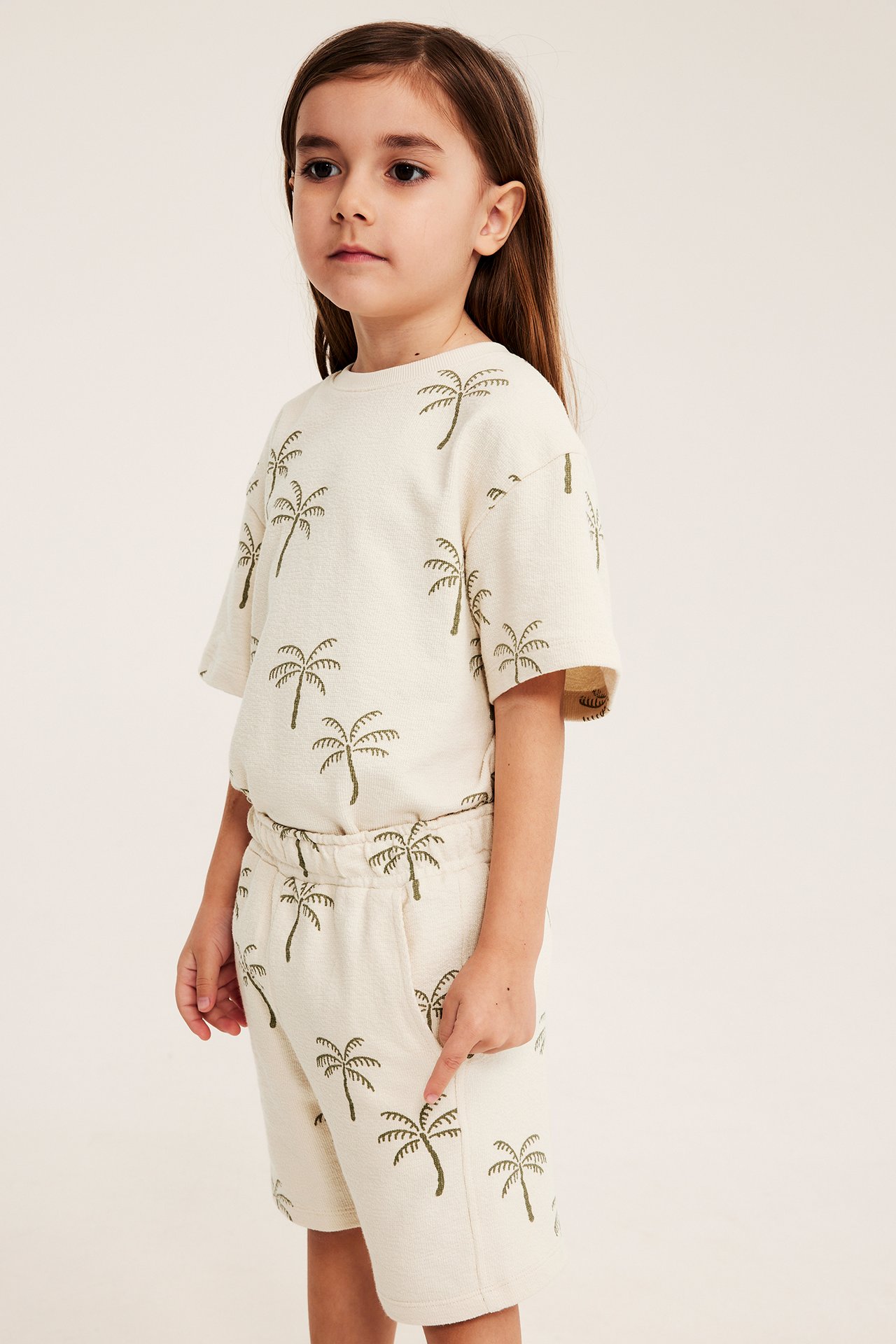 Frontal sidovy: barn i ljusbeige jersey-shorts med fickor och matchande t-shirt med palmprint.