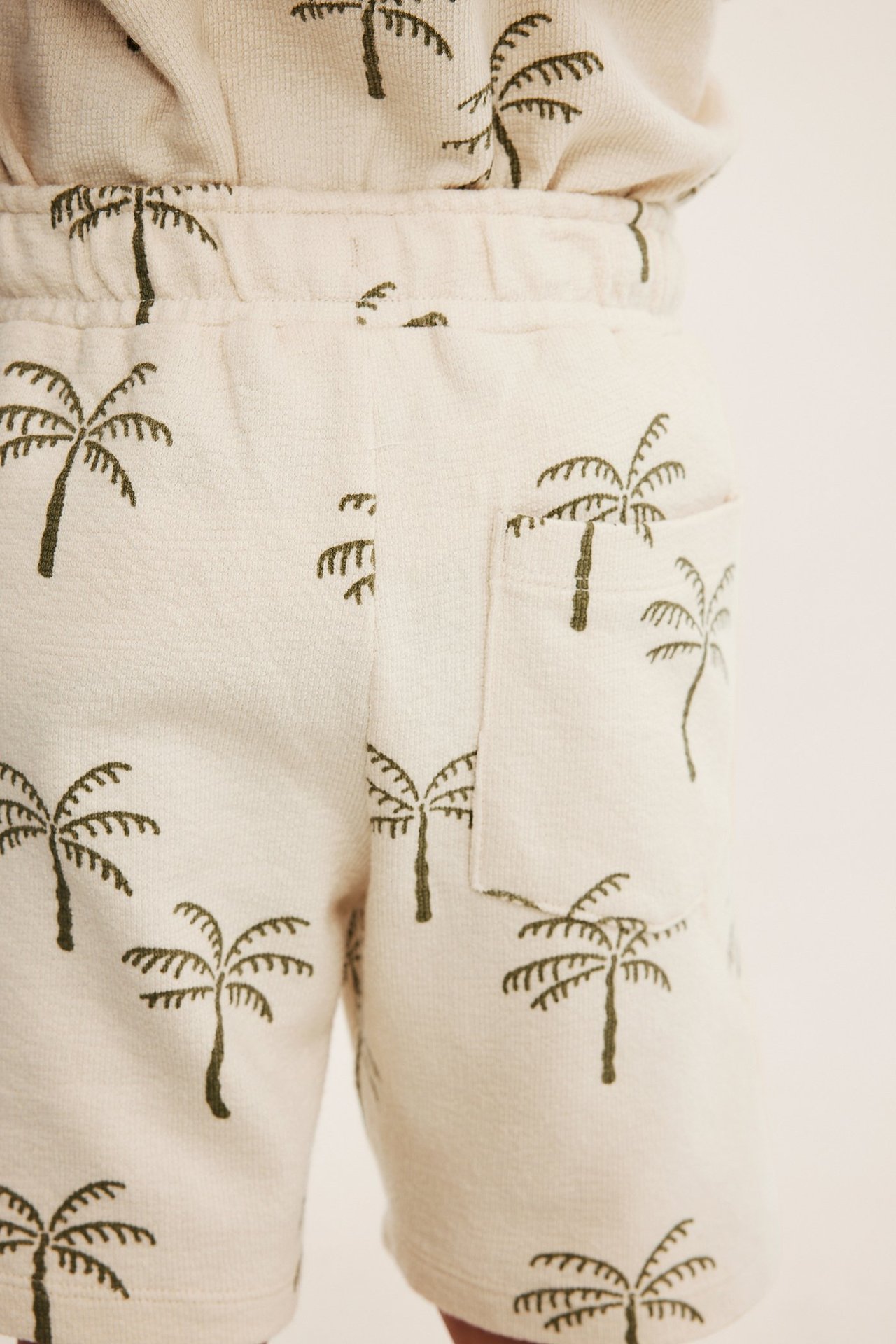 Lähikuva lasten vaaleabeigeistä puuvillajerseyshortseista, joissa tummanvihreä palmukuvio ja sivutasku.