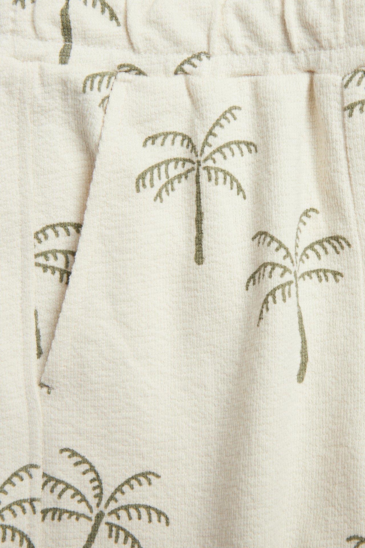 Närbild av ljusbeige bomullstrikåshorts med grönt palmönster, visar ficka och resår i midjan.
