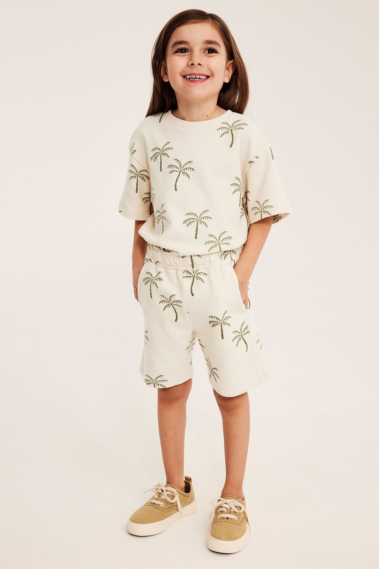 Lapsi edestä: vaaleanbeiget kuvioidut jersey-shortsit, yhteensopiva t-paita ja ruskeat lenkkarit.