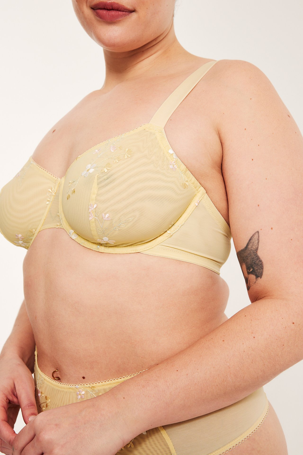 Sidebilde av kvinne i lysgul spile-BH i mesh med broderte blomster og matchende truse.