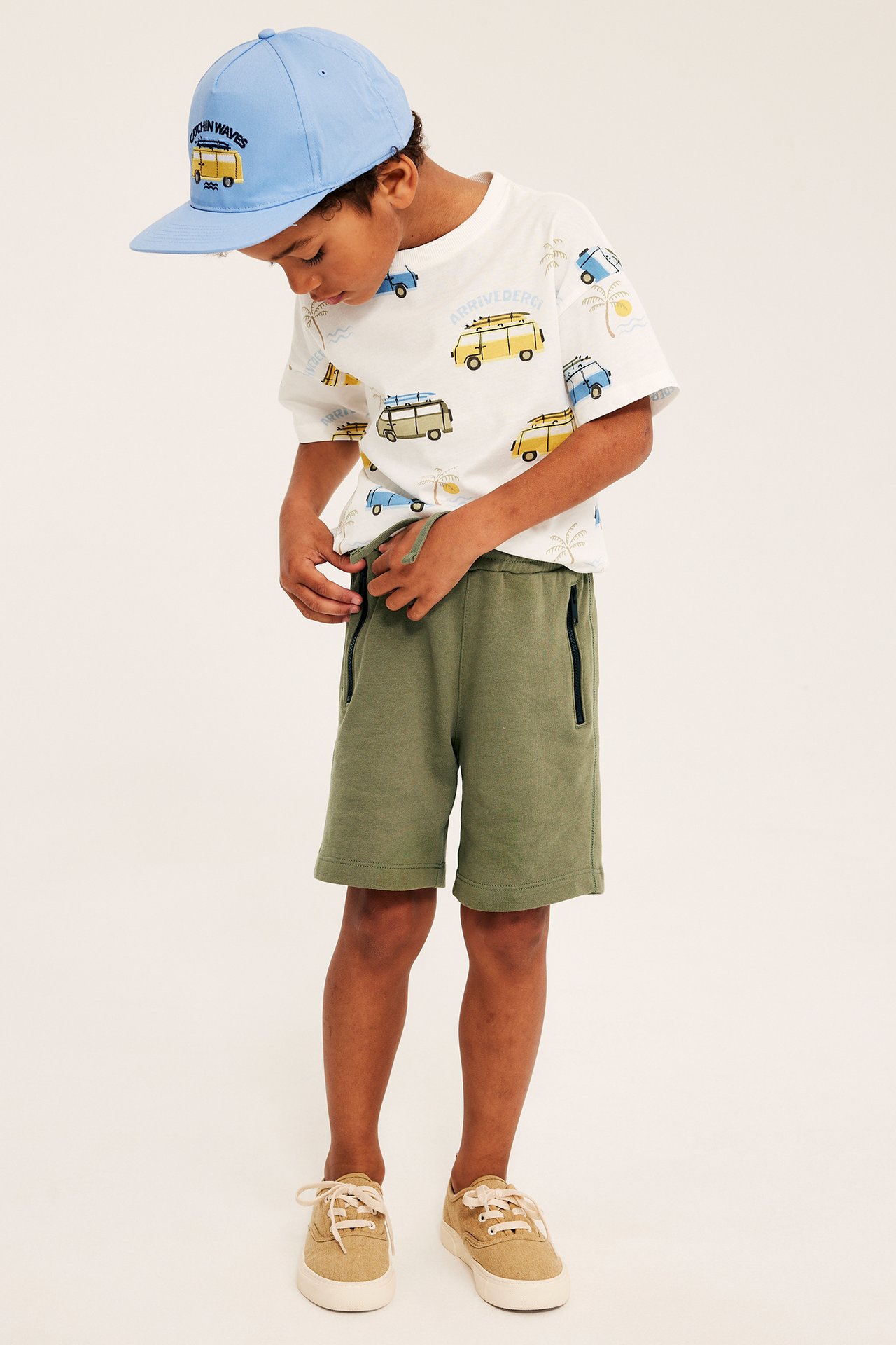 Barn bär mörkgröna mjukisshorts med dragkedjefickor, mönstrad t-shirt, ljusblå keps och beige sneakers.