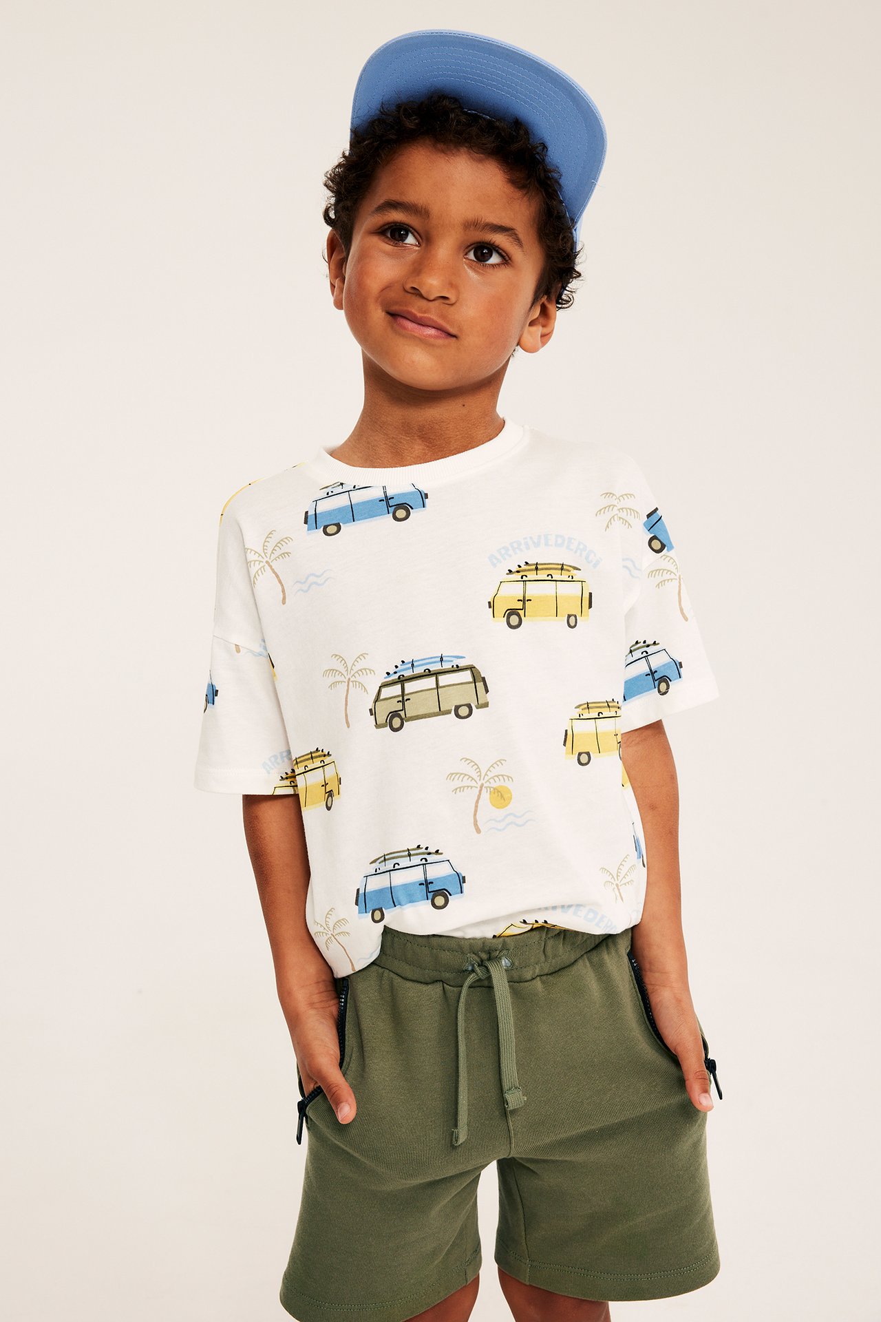 Edestä: lapsi tummanvihreissä verryttelyshortseissa vetoketjutaskuilla, valkoinen kuvioitu t-paita ja sininen lippalakki.