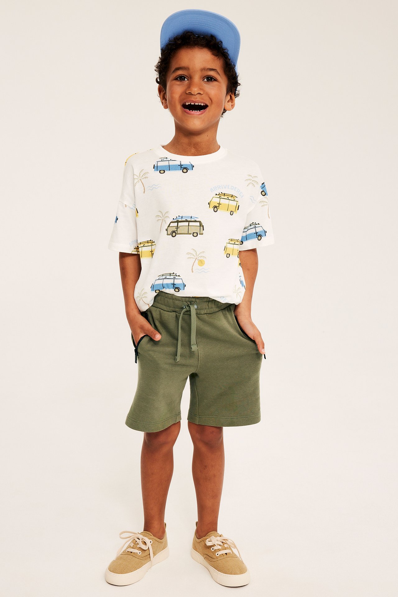 Framifrån, en pojke i mörkgröna jerseyshorts med dragsko, vit mönstrad t-shirt, blå keps och sneakers.