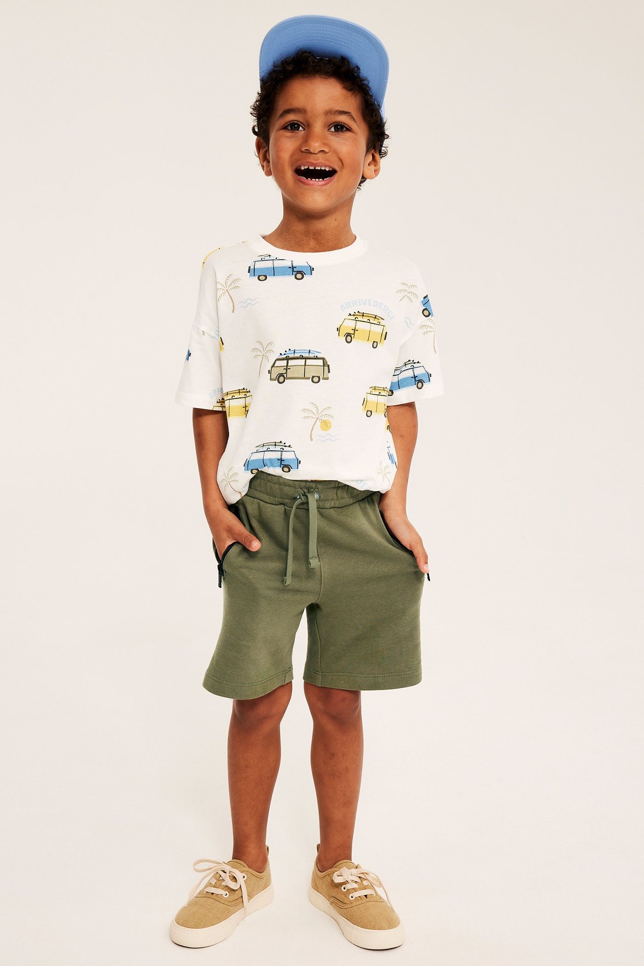 Edestäpäin poika, yllään tummanvihreät collegeshorts kiristysnyörillä, valkoinen painettu t-paita, sininen lippalakki ja lenkkarit.