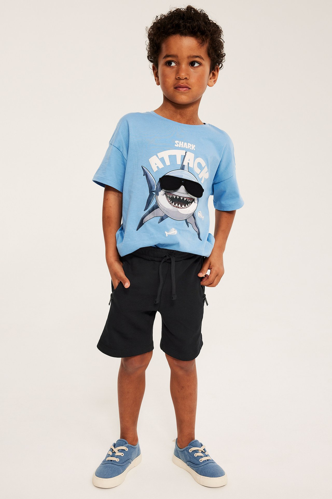 Frontvy av ett barn i svarta mjukisshorts med dragsko, blå t-shirt med hajmönster och blå tygskor.