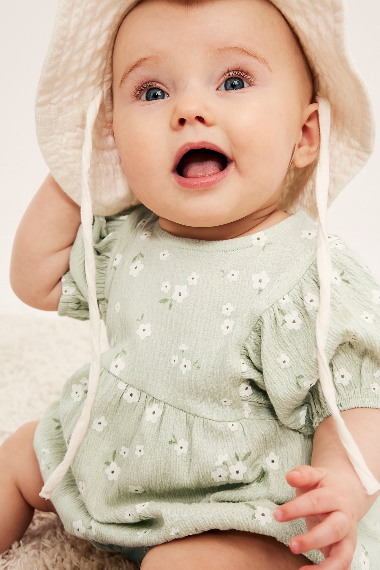 Nærbilde, forfra, av baby i lysegrønn blomstret romper med puffermer og offwhite bøttehatt.