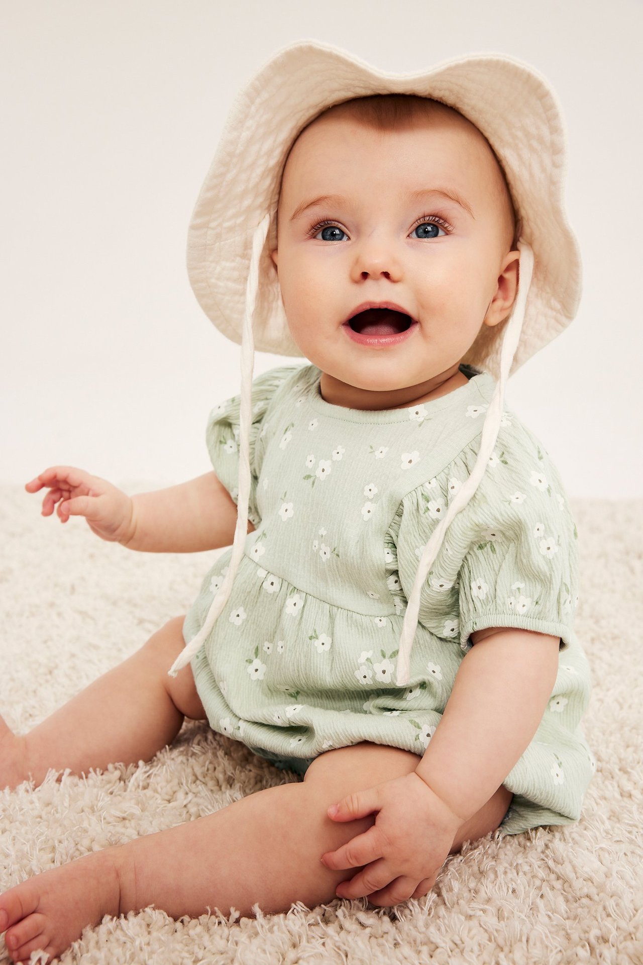 Vauva edestä: yllään vaaleanvihreä kukkakuvioinen pussuhihainen romper ja valkoinen hattu.