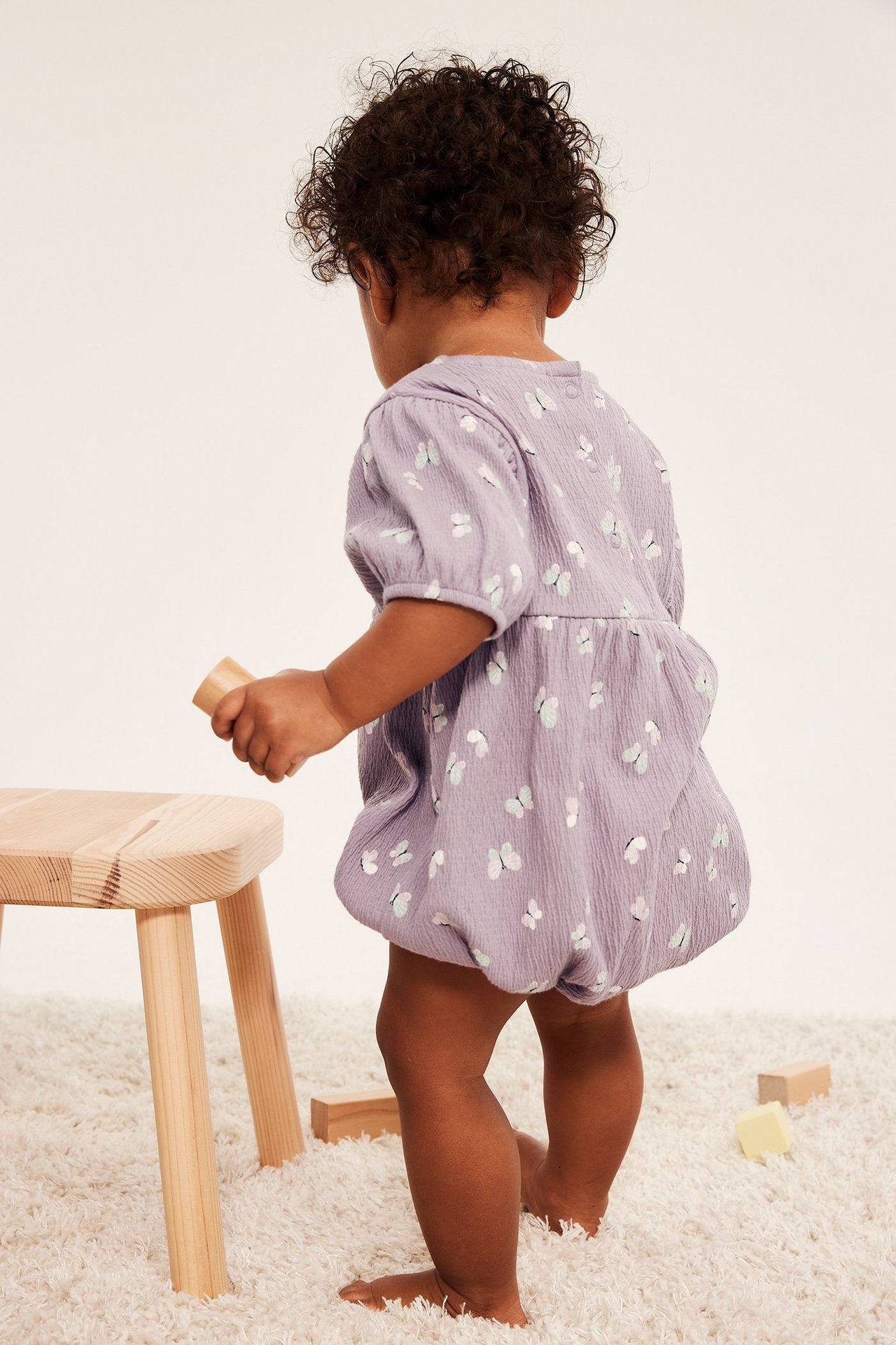 Vauvan selkäpuoli lilaa romper-haalaria, jossa pöyhkeät hihat ja perhoskuvio, pitäen puupalikkaa.