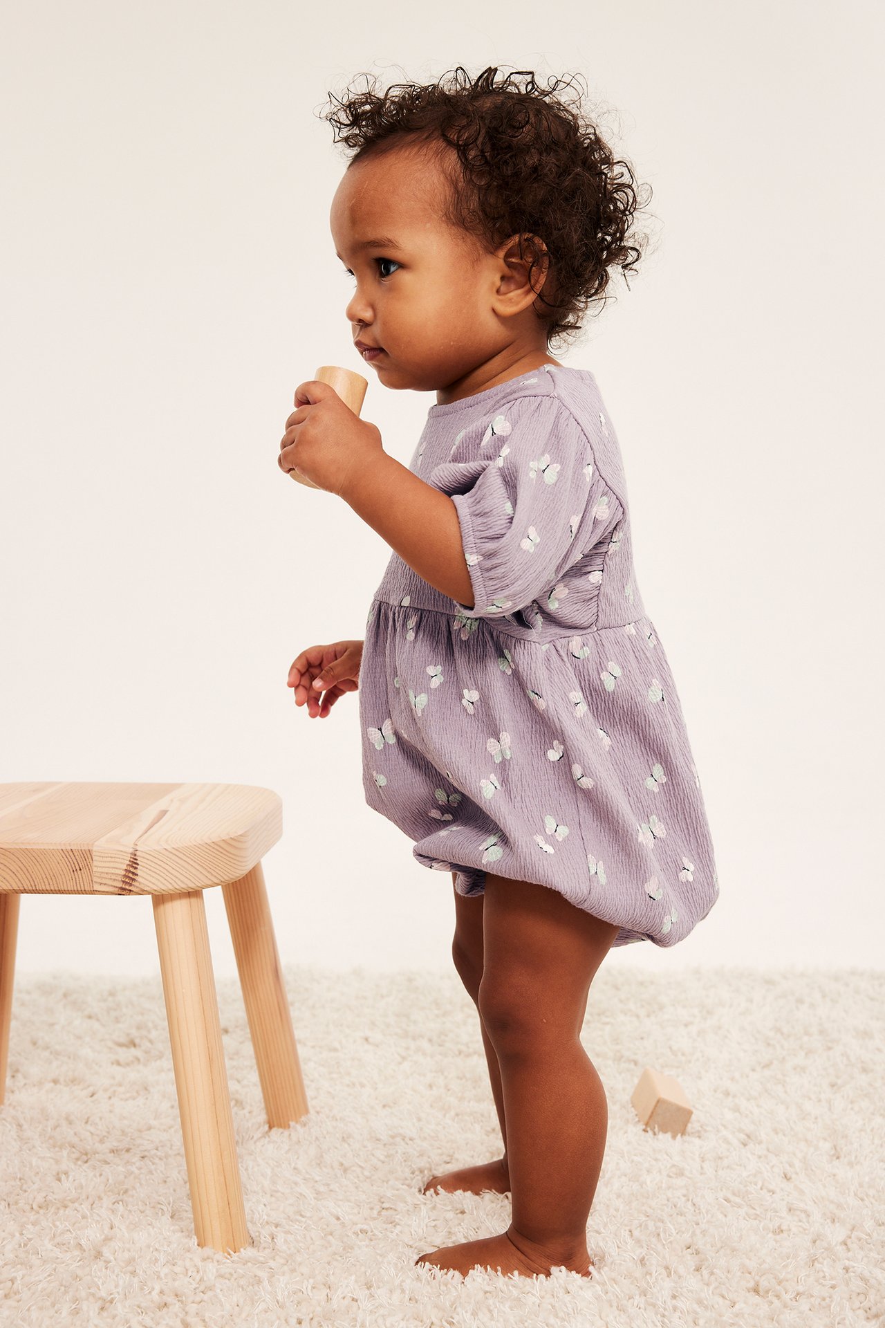 Sivukuva vauvasta lila-kuvioidussa perhosromperissa, jossa pussihihat, pidellen puupalikkaa.