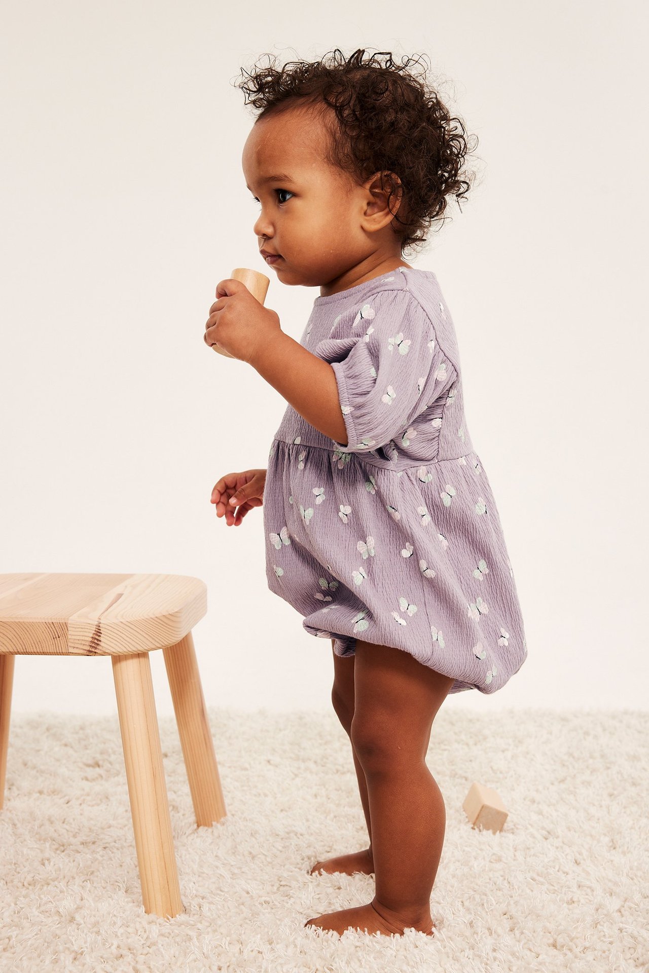 Widok z boku niemowlęcia w liliowym romperze w motyle z bufiastymi rękawami, trzymającego drewniany klocek.