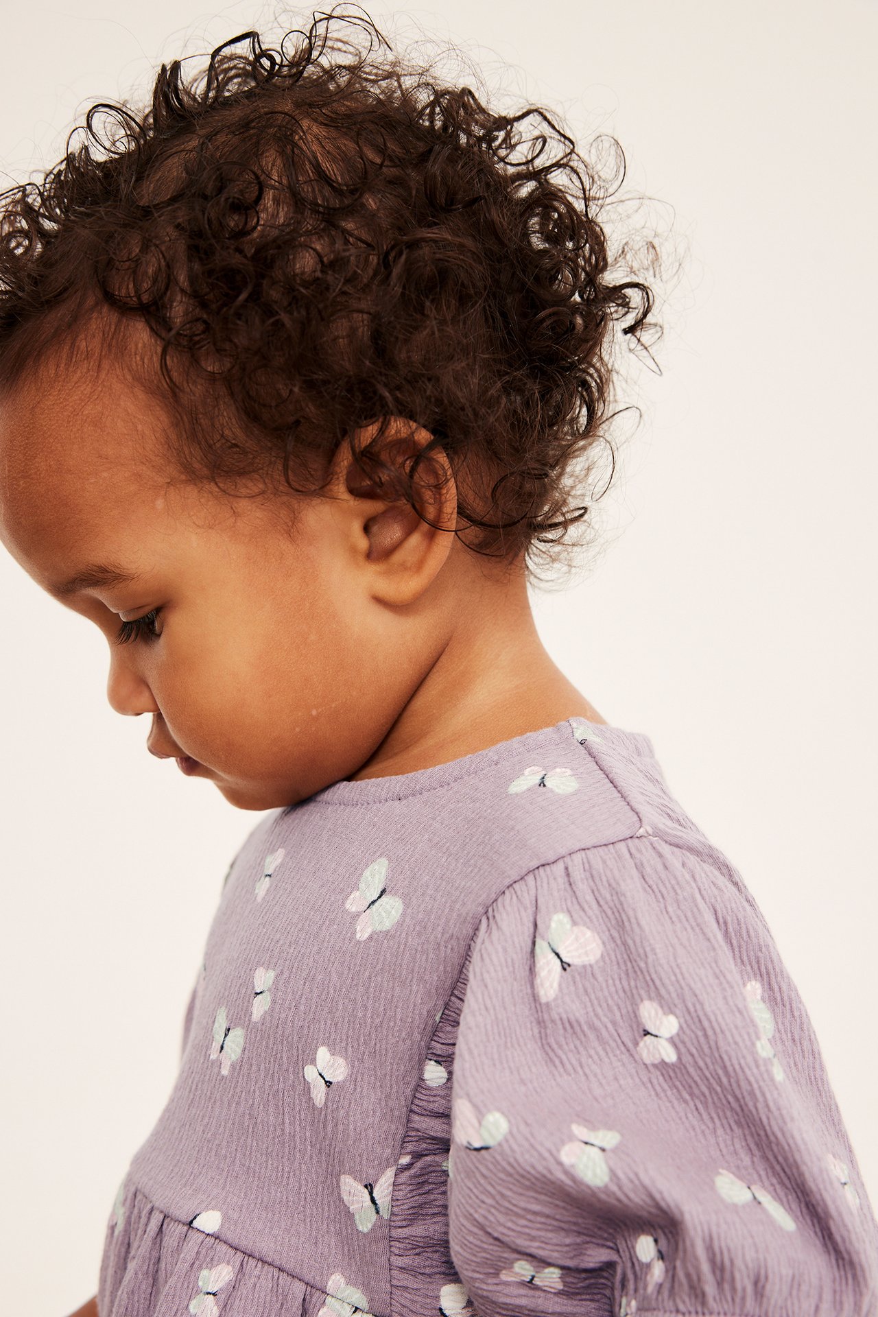 Lähikuva sivukuva vauvasta, yllään lila teksturoitu romper puhvihihoilla ja perhoskuvioinnilla.
