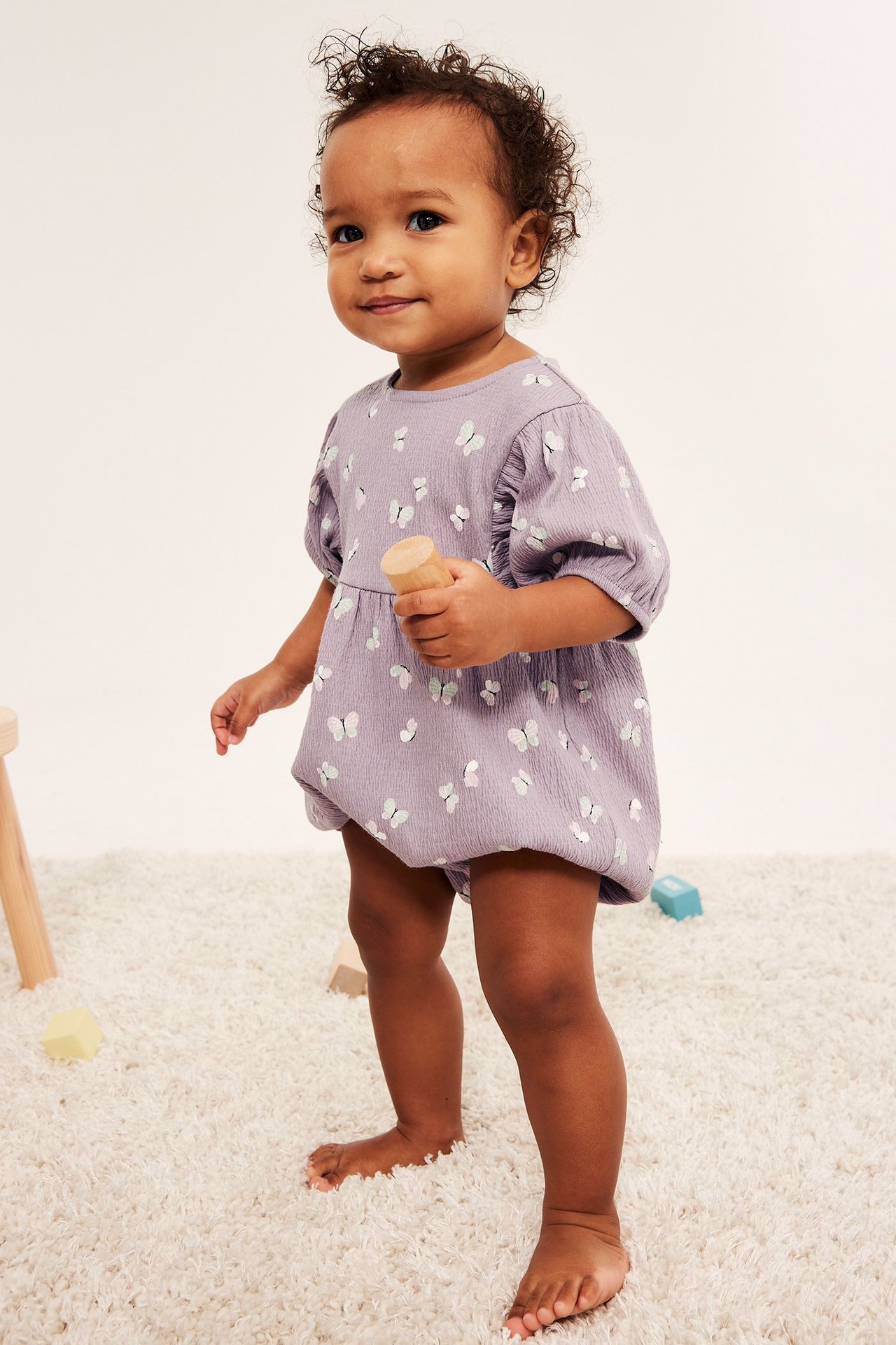Edestäpäin: Vauva yllään lila, perhoskuvioitu, pussihihainen potkupuku, pitää puulelua.