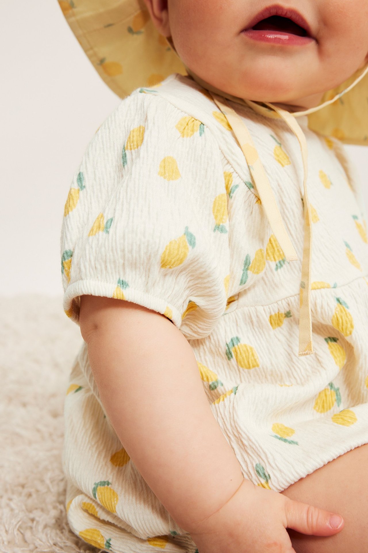 Närbild på baby i offwhite citronmönstrad romper med puffärmar och matchande solhatt.
