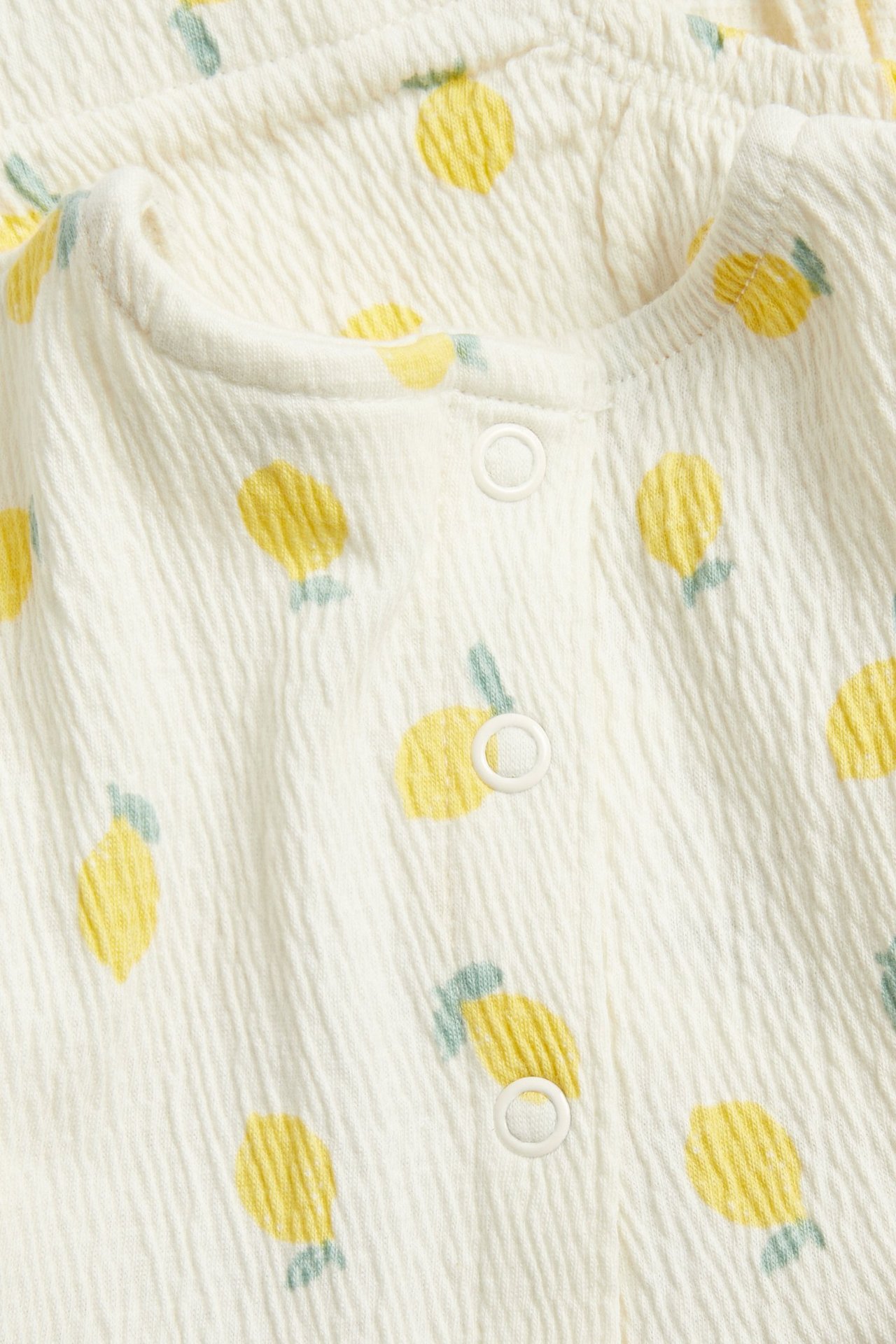 Lähikuva luonnonvalkoisesta vauvan romperista, jossa teksturoitu kangas, keltainen sitruunakuvio ja valkoiset nepparit.