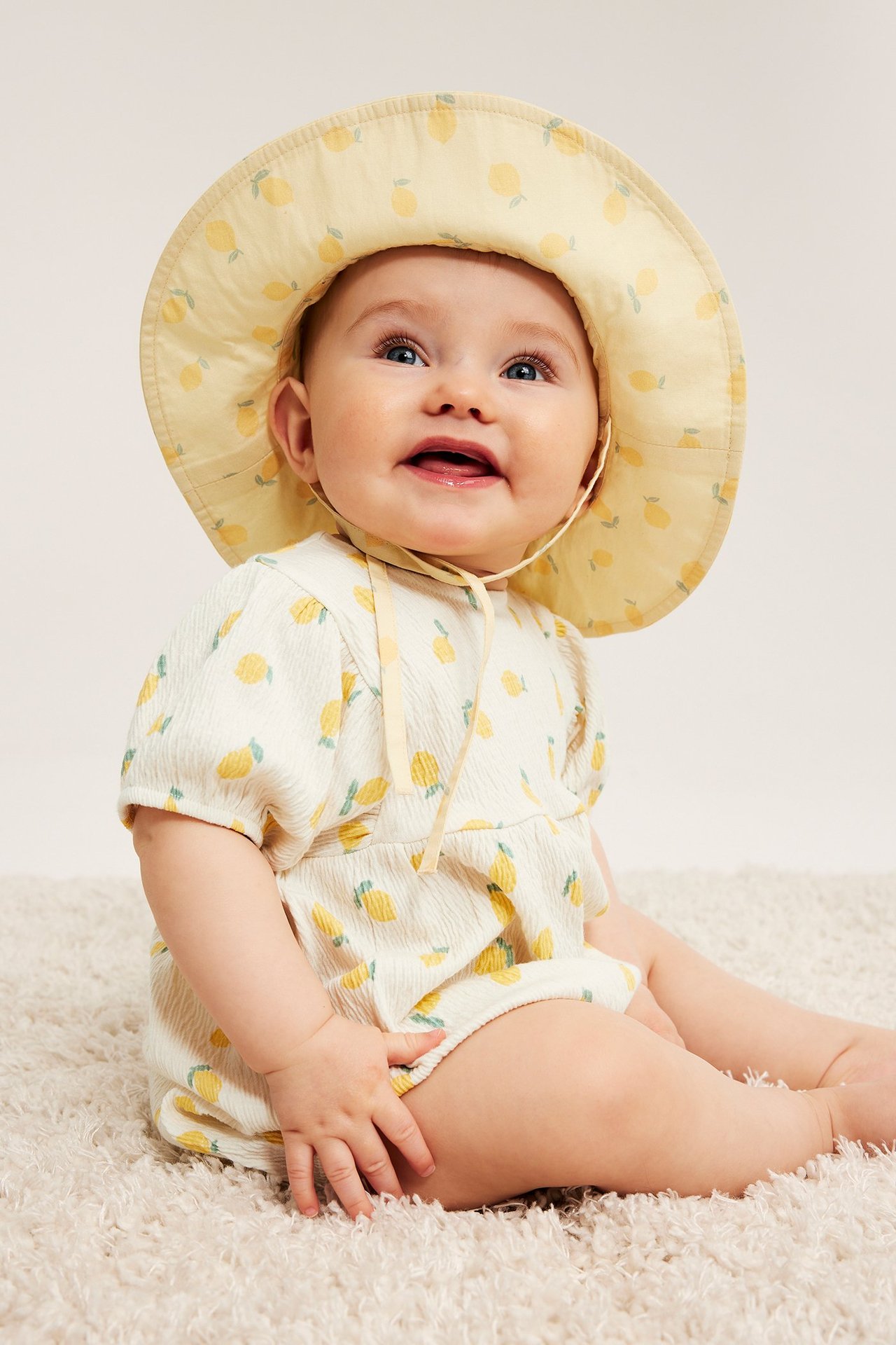 Framifrån, en baby i en offwhite kortärmad romper med citronmönster och en gul citronmönstrad solhatt.