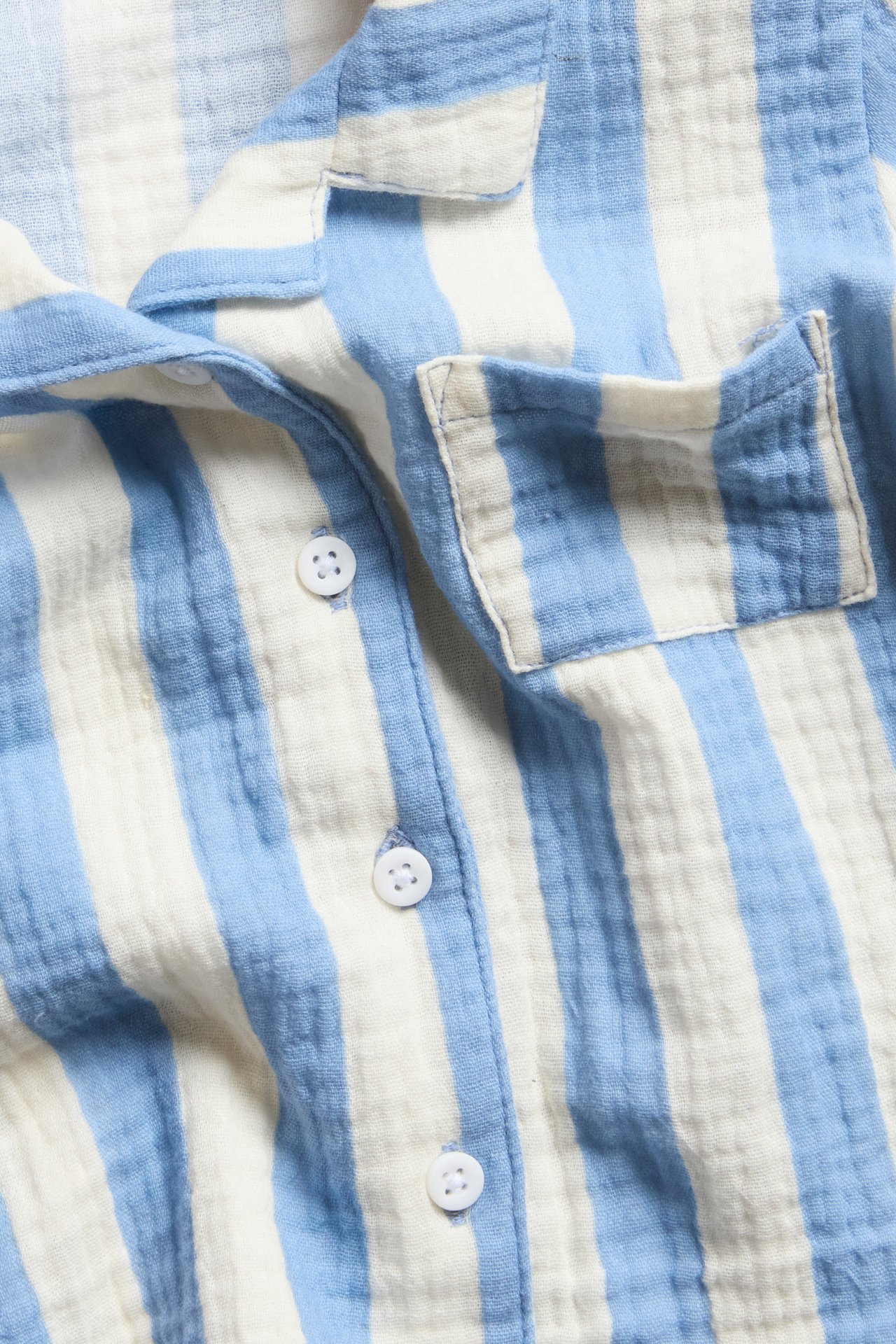 Lähikuva vaaleansinisestä ja valkoisesta raidallisesta musliinipaidasta, jossa rintatasku ja valkoiset napit.