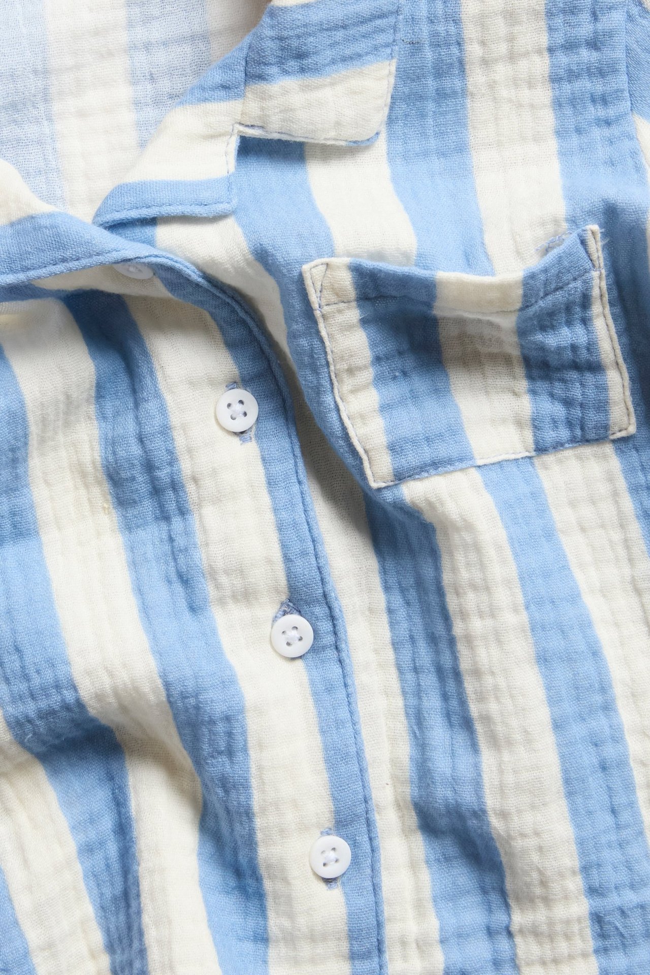 Zbliżenie na jasnoniebiesko-białą koszulę muślinową w paski, z kieszonką na piersi i białymi guzikami.