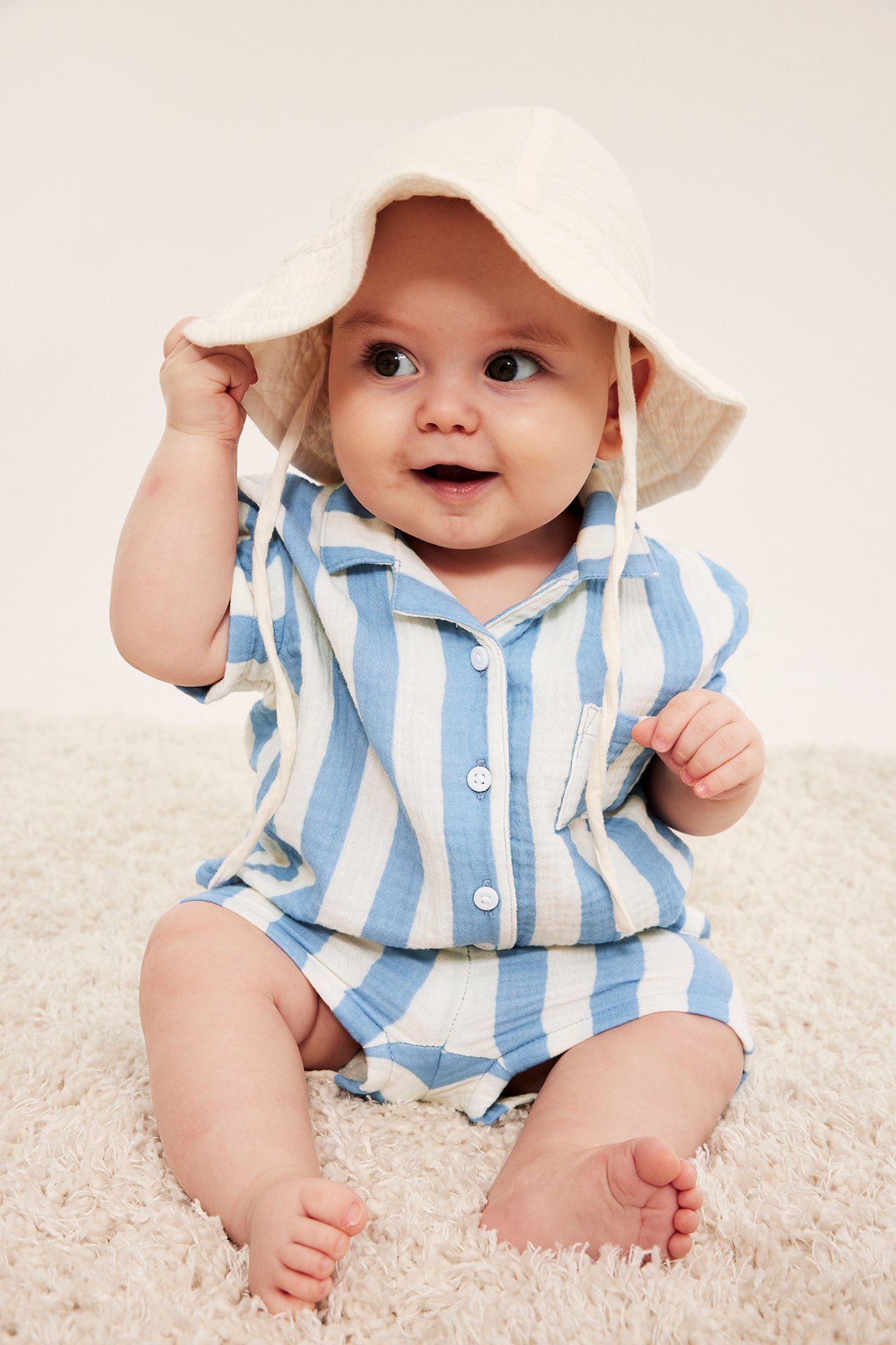 Framifrån: en baby i ljusblårandig muslin skjorta, matchande shorts, och en benvit solhatt.