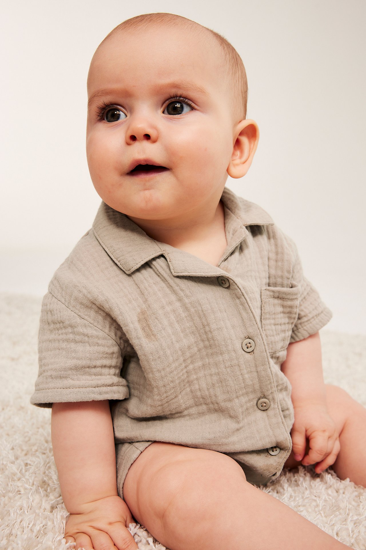 Frontal-sidebilde: baby i en beige kortermet bomullsmuslin-skjorte med knapper og matchende underdel.