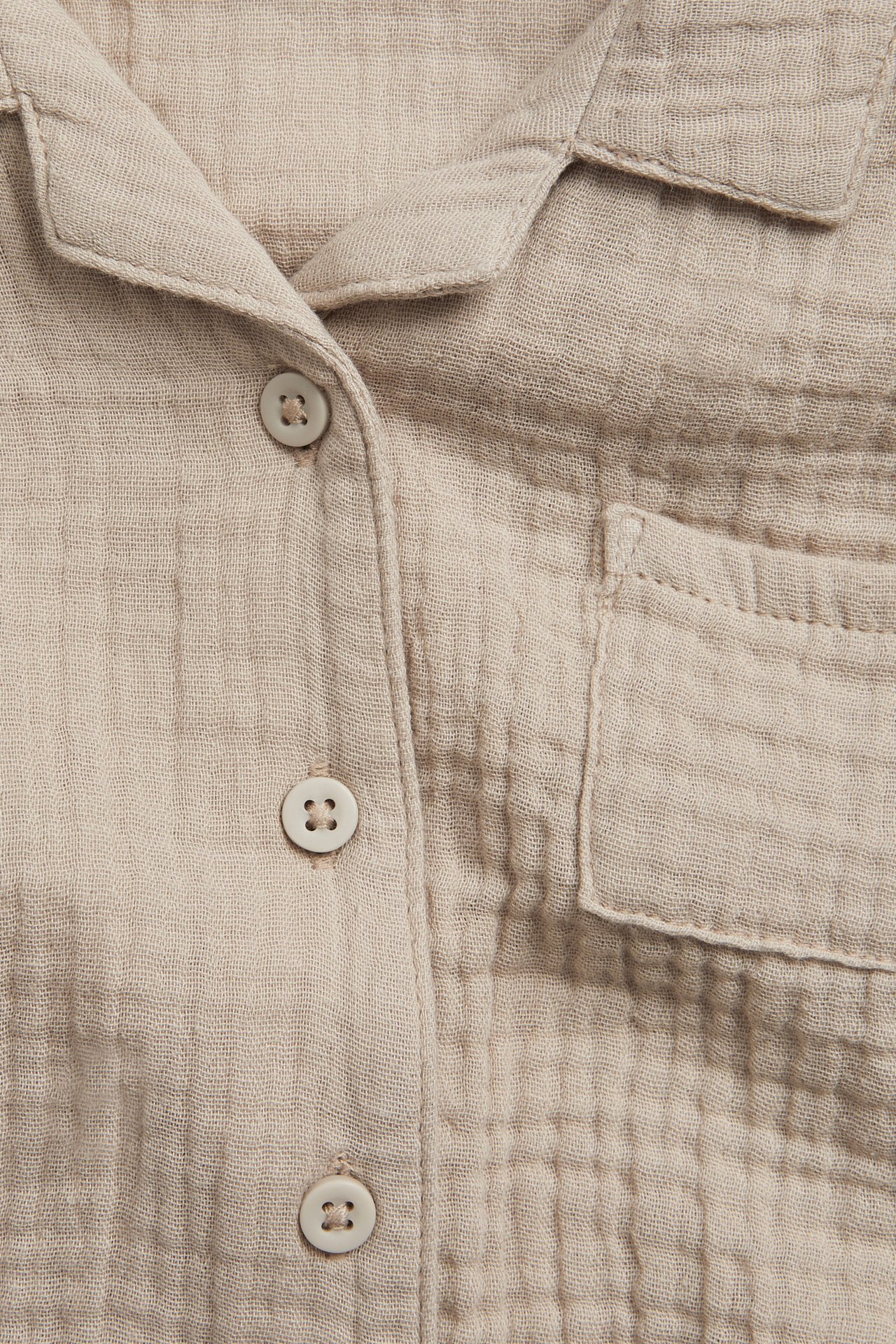 Lähikuva edestä beigestä lyhythihaisesta musliinipaidasta, jossa napit ja rintatasku.