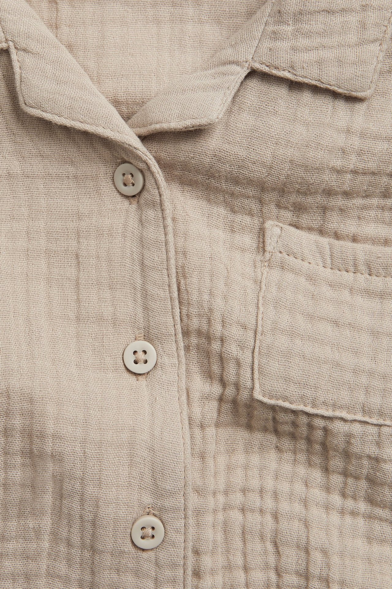 Zbliżenie z przodu beżowej koszuli muślinowej z krótkim rękawem, z guzikami i kieszenią na piersi.