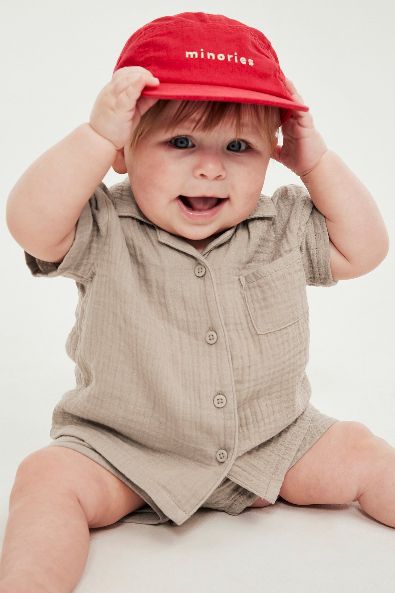 Edestä päin: Vauva yllään beige musliininen lyhythihainen paita napeilla, yhteensopivat shortsit ja punainen lippalakki.