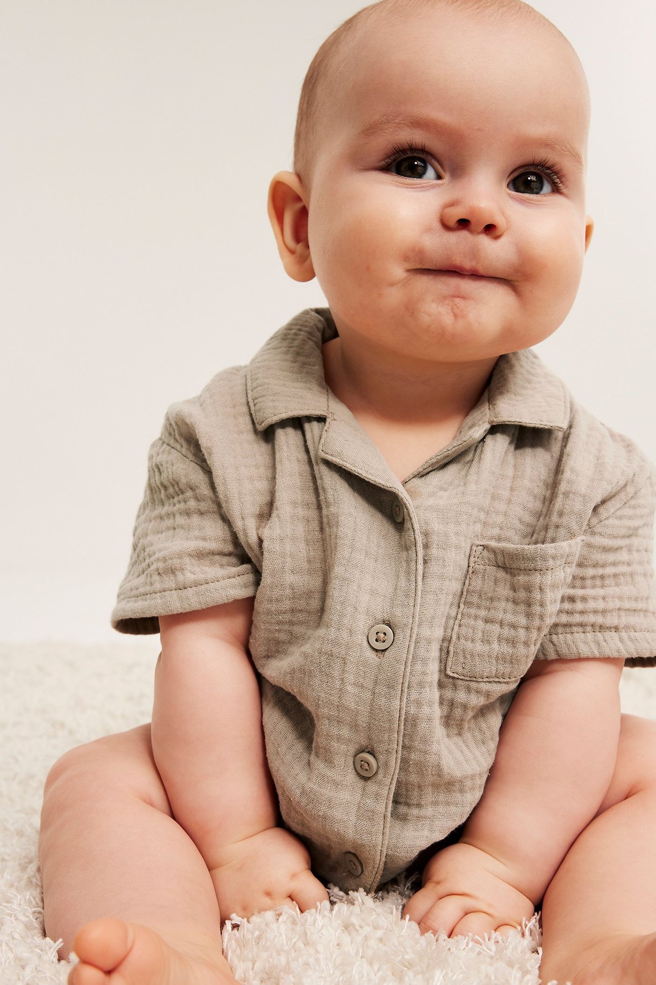 Zbliżenie, przód: dziecko w beżowej, krótko rękawowej koszuli muślinowej z guzikami i kieszenią na piersi.