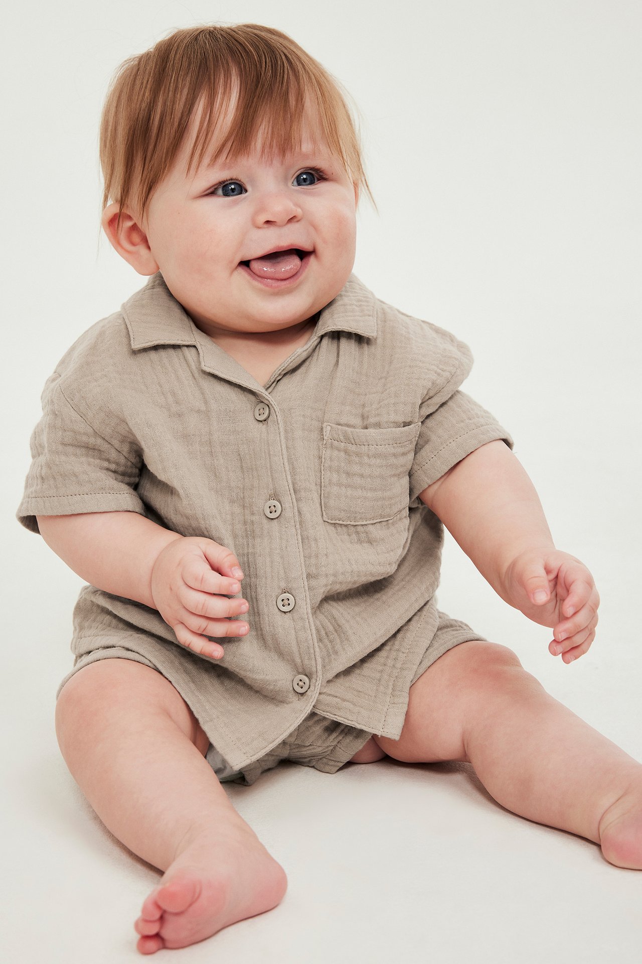 Framifrån, en leende baby i beige kortärmad muslin skjorta med ficka och matchande shorts.