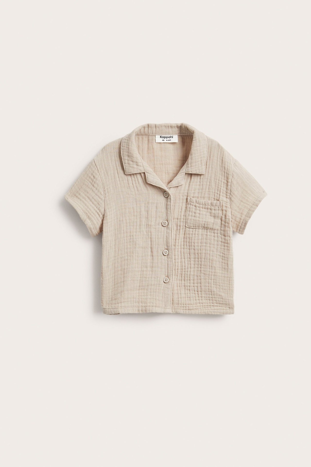 Edestä näkymä beigestä lyhythihaisesta musliinipaidasta, jossa kaulus, nappikiinnitys ja rintatasku.