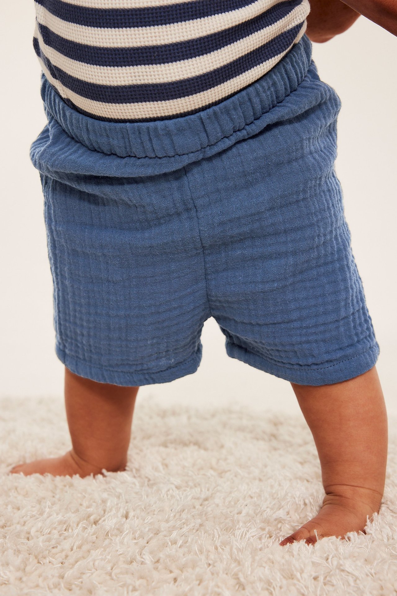 Bakifrån, en baby bär blå muslinshorts med resår i midjan och en marinblå och vitrandig topp.