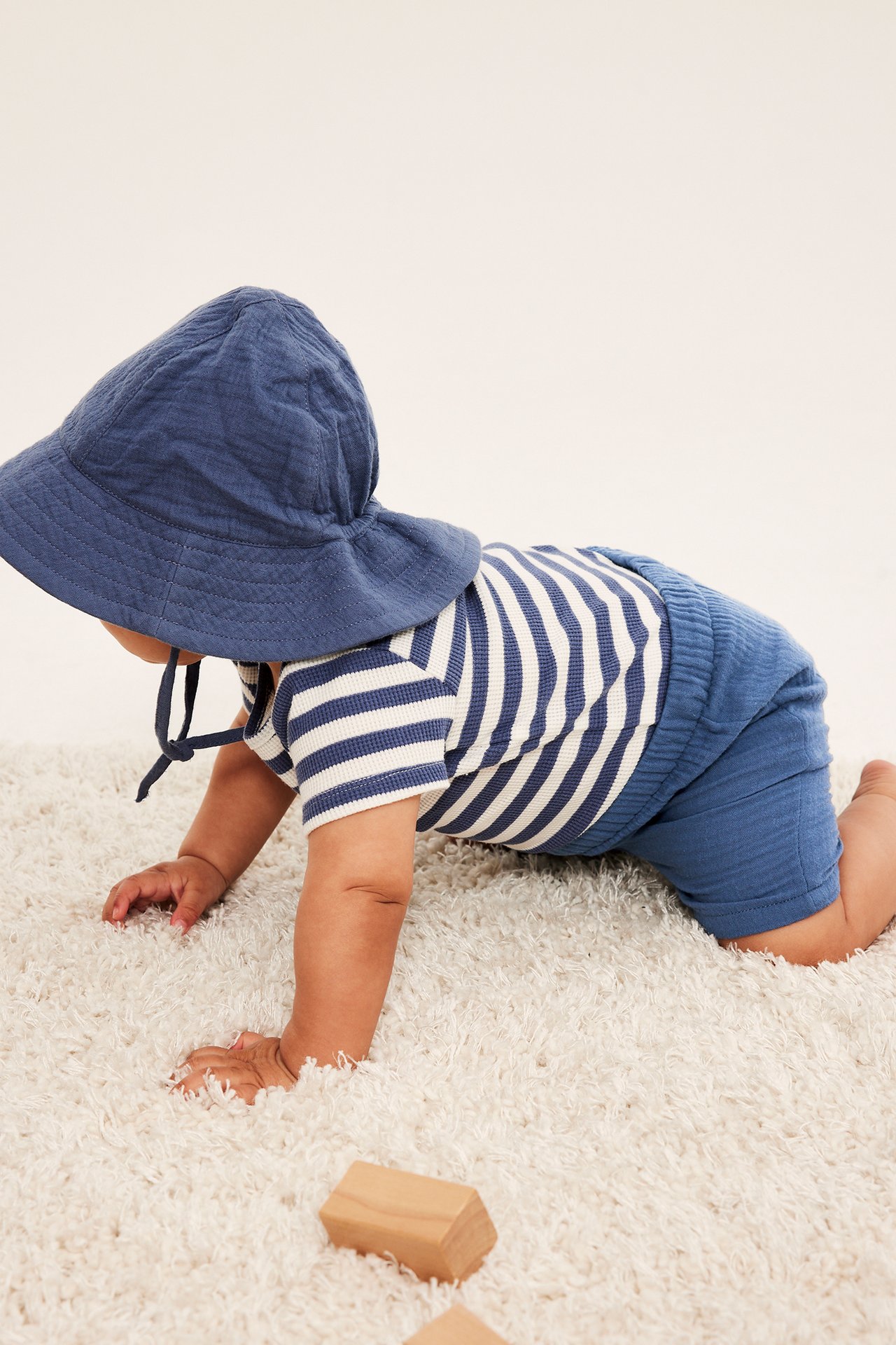 Sidevisning: Baby kryper i blå muslinshorts, en stripete topp og en matchende blå muslinbøttehatt.