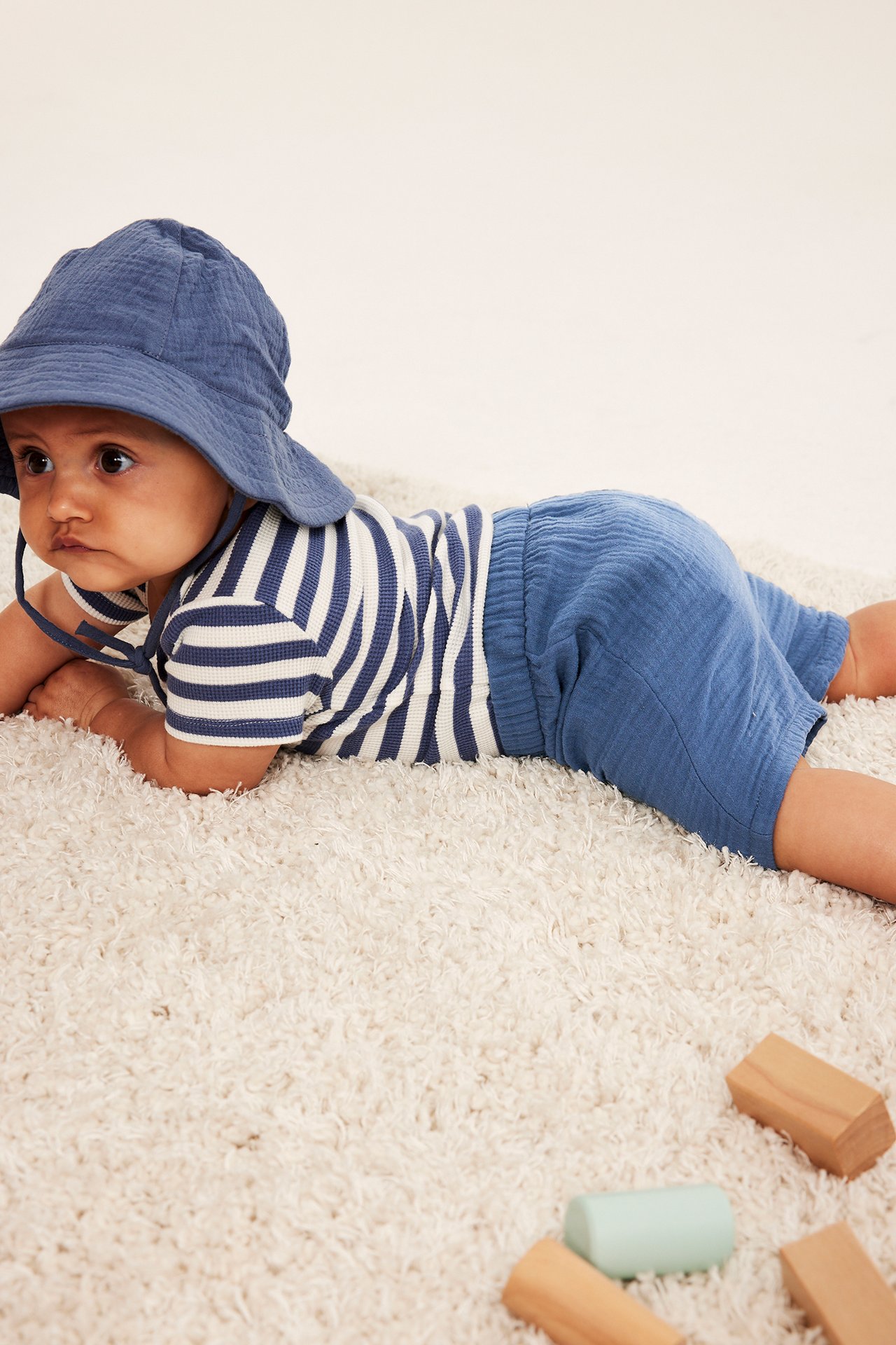 Widok z boku: niemowlę w niebieskich muślinowych szortach z gumką, pasiastym T-shircie i niebieskim kapeluszu.