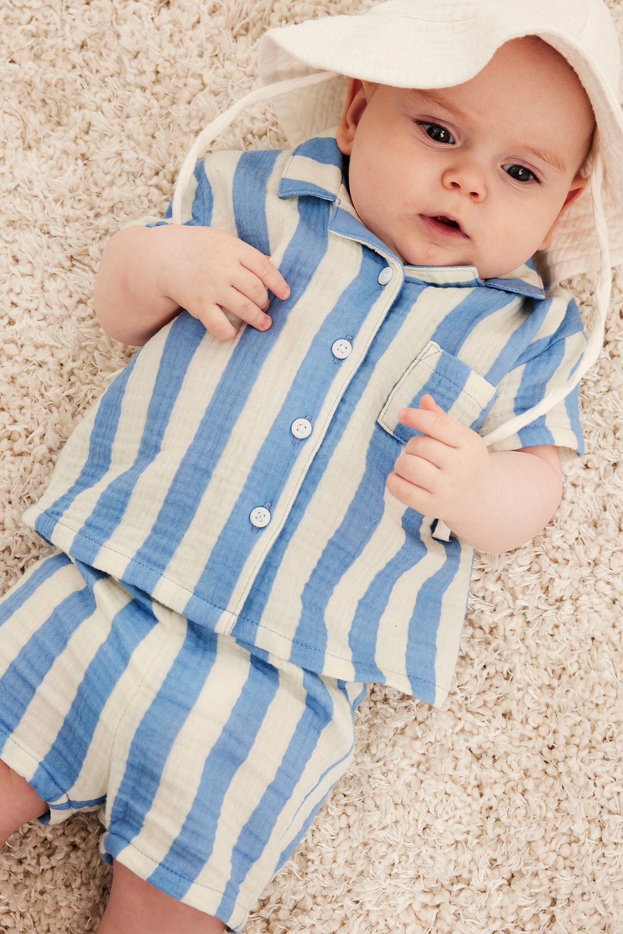 Baby bär ljusblå randiga muslinsorts, matchande skjorta och vit solhatt. Framifrån sett.