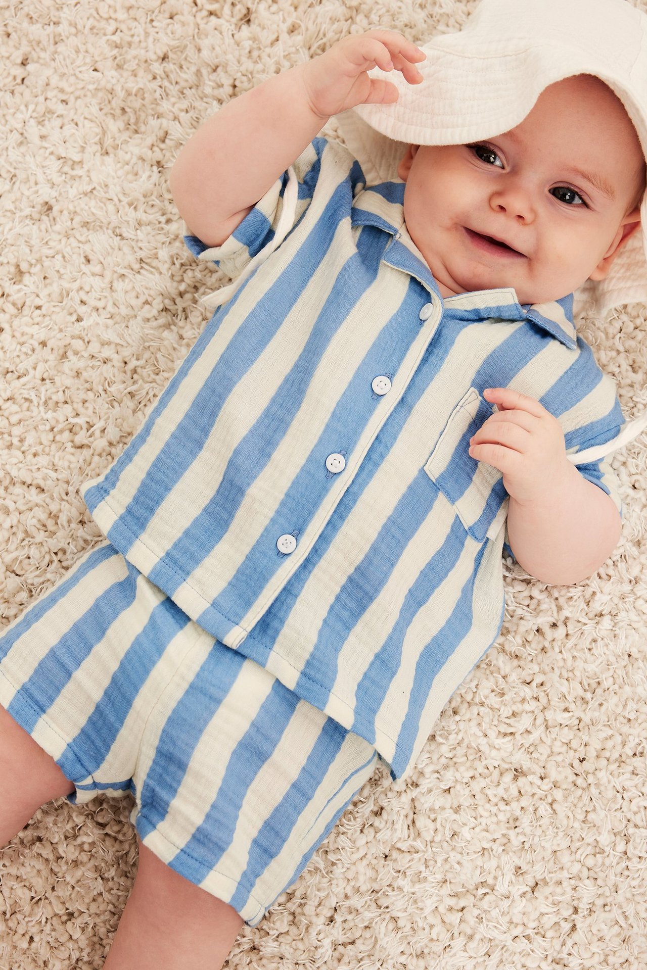 Fågelperspektiv av baby i ljusblårandiga muslinshorts, matchande skjorta och vit hatt.