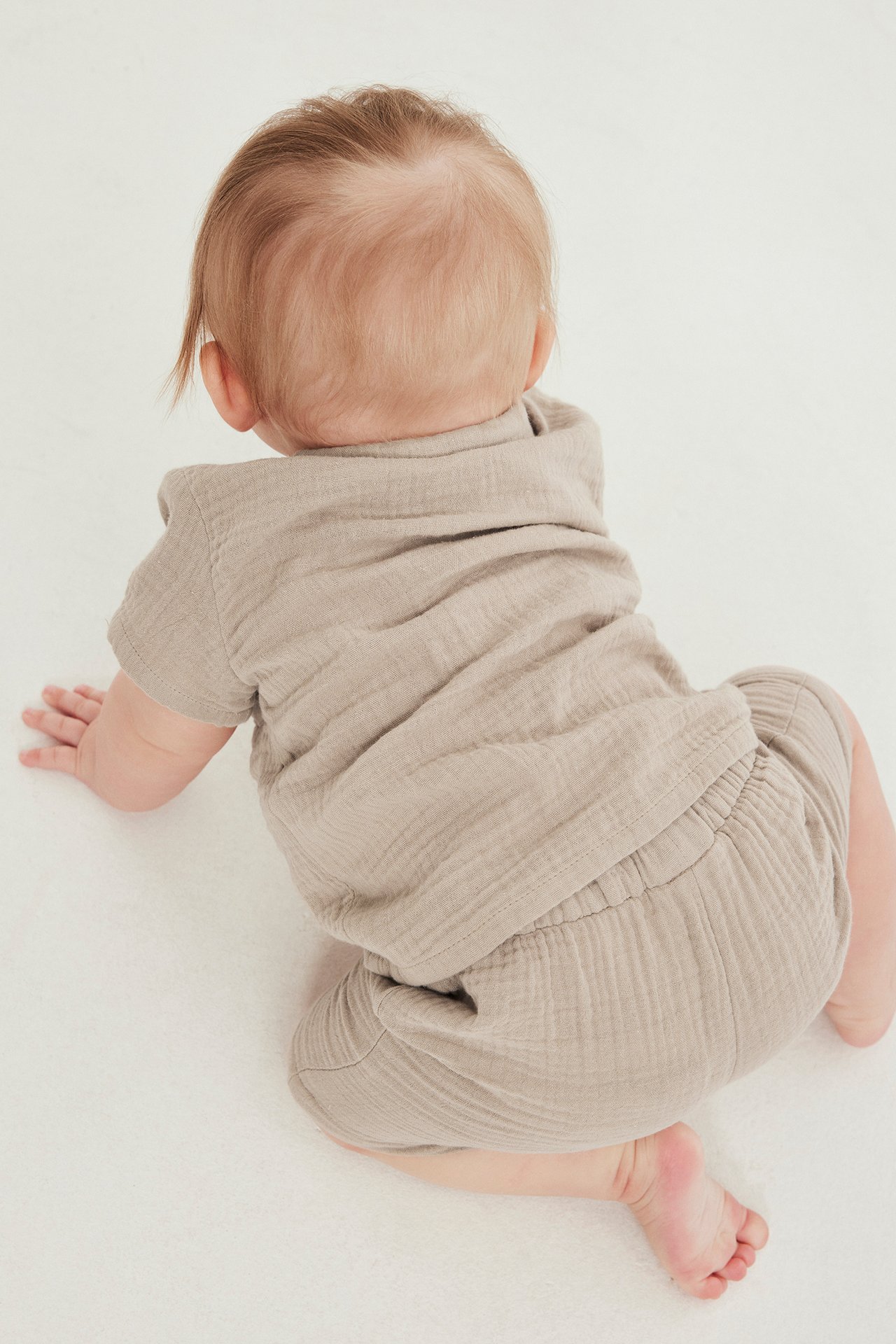 Takaapäin näkymä vauvasta ryömimässä, yllään beiget harsoshortsit joustavalla vyötäröllä ja yhteensopiva toppi.