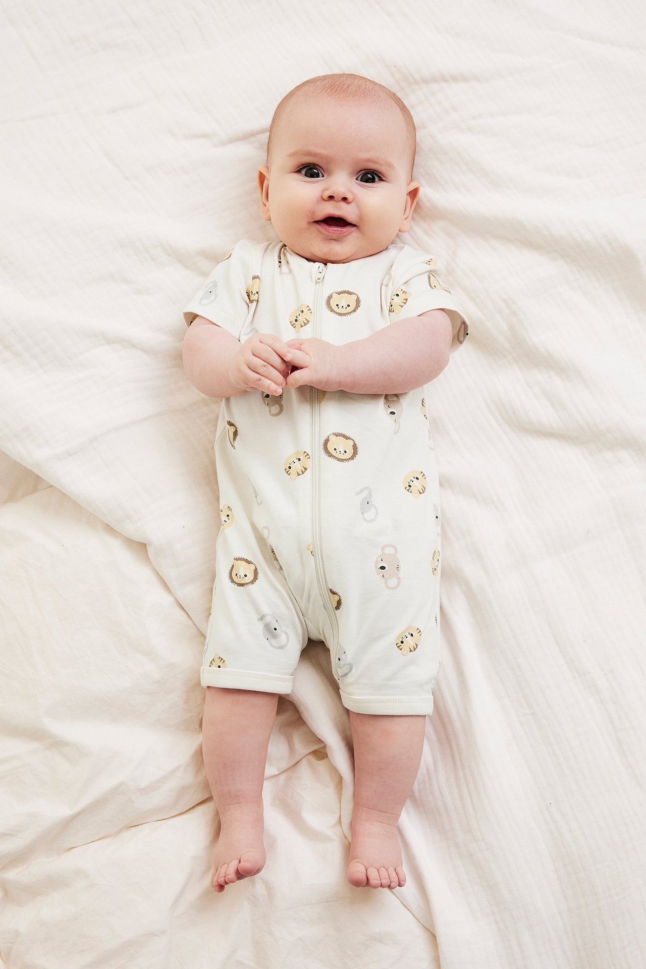 Widok z góry: niemowlę w ecru kombinezonie z krótkim rękawem, zwierzęcym wzorem i zamkiem z przodu.