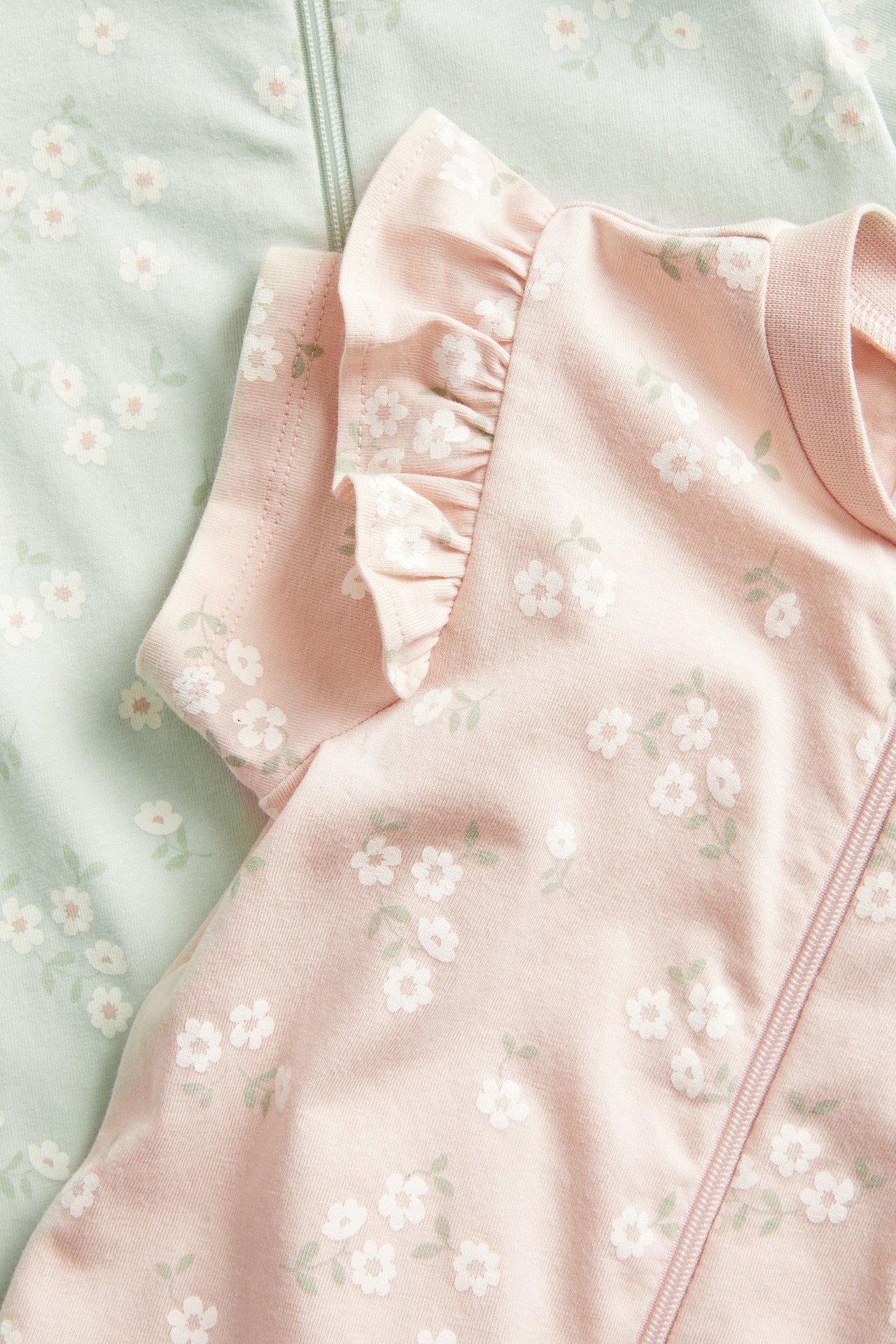 Närbild av en rosa kortärmad babyromper med volanger och blommigt tryck, bredvid en mintgrön romper.