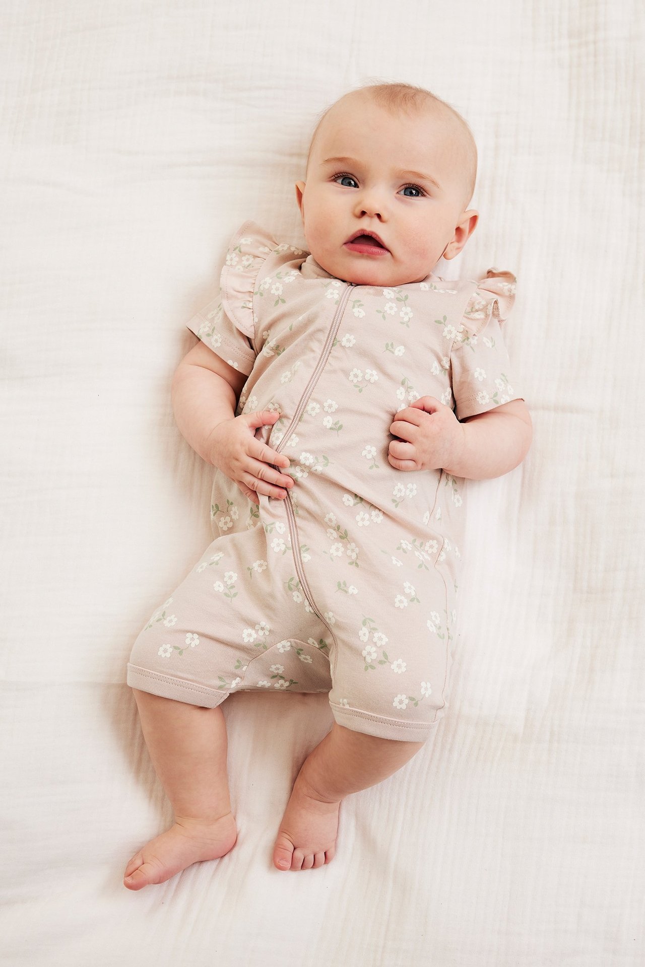 Widok z przodu: niemowlę w różowym, kwiecistym kombinezonie z krótkim rękawem, falbanami i zamkiem.