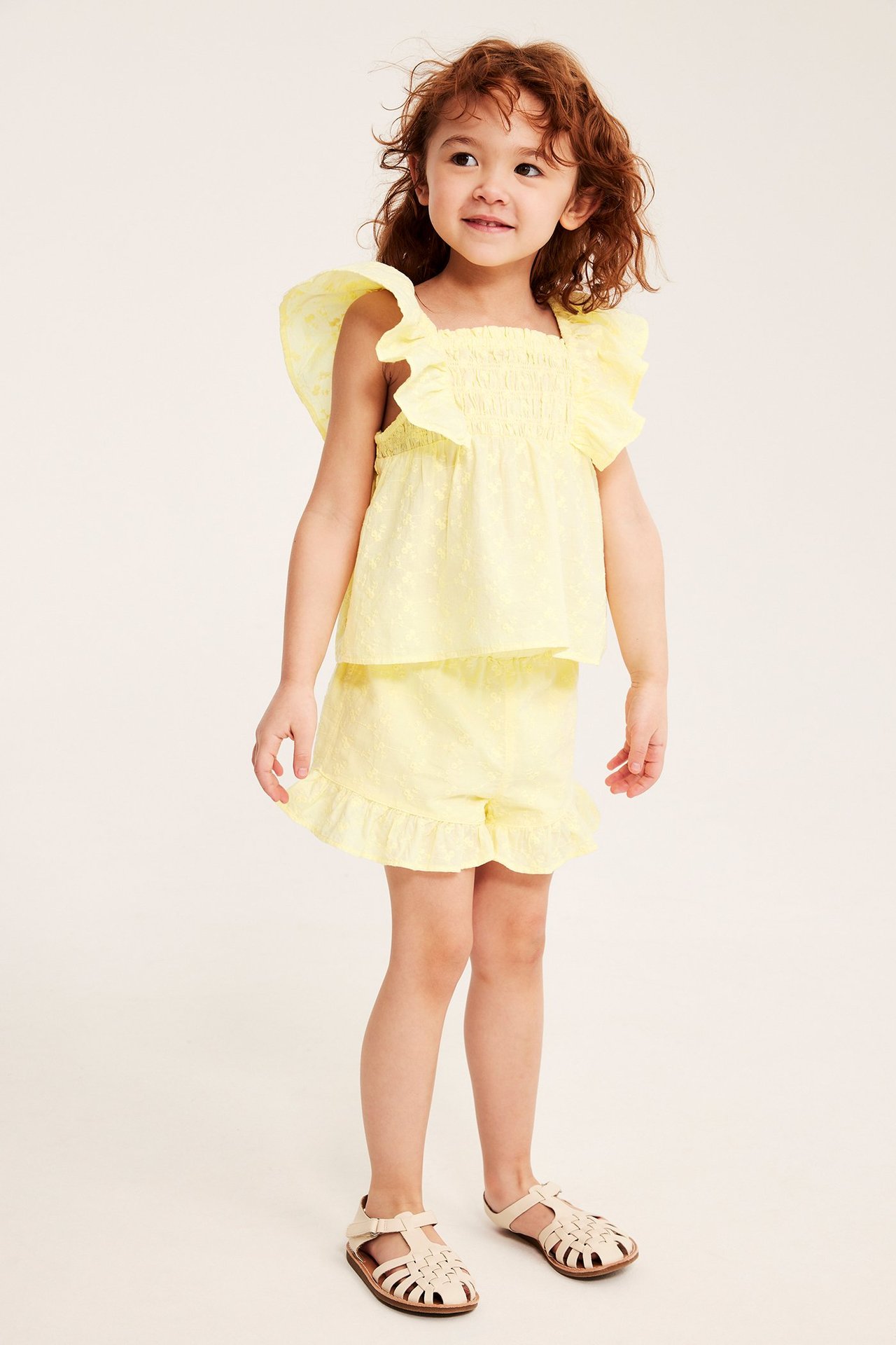 Framifrån, ett barn i ljusgul broderad volangblus, matchande shorts och ljusbeige sandaler.