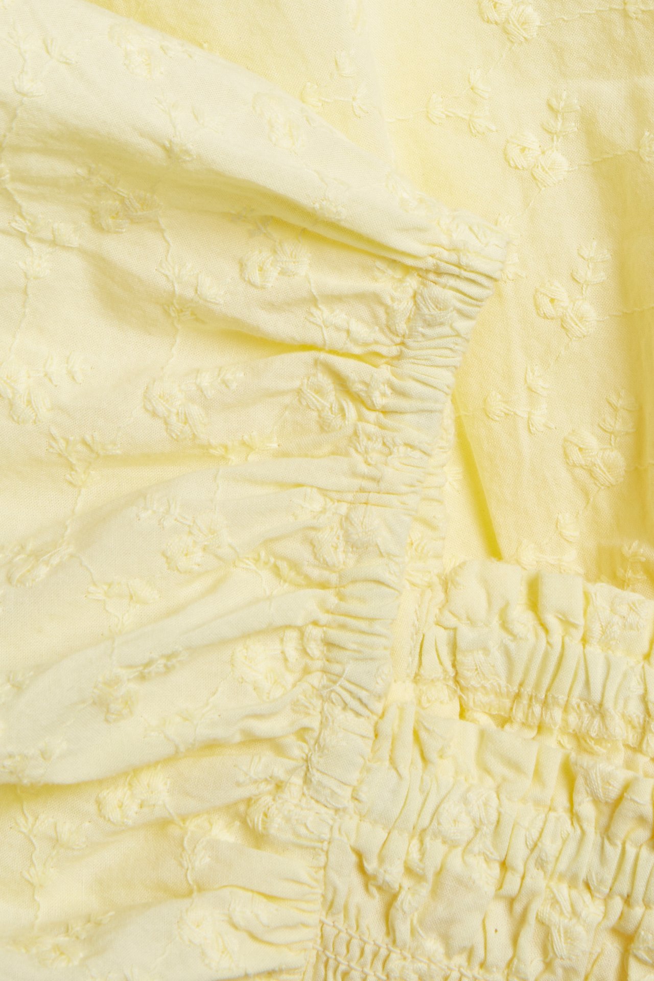 Närbild av en ljusgul volangblus i bomull med broderade blommönster och rynkade detaljer.