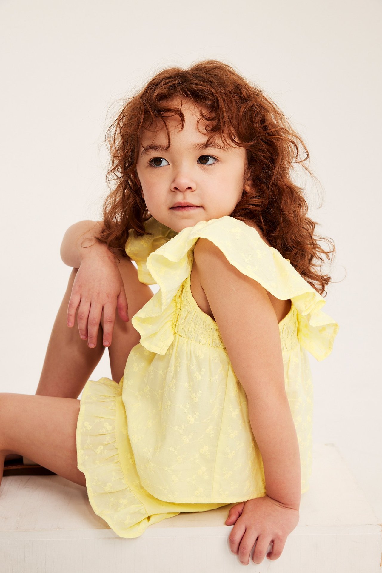 Framsida av ett sittande barn i en ljusgul volangblus i bomull med brodyr och matchande shorts.