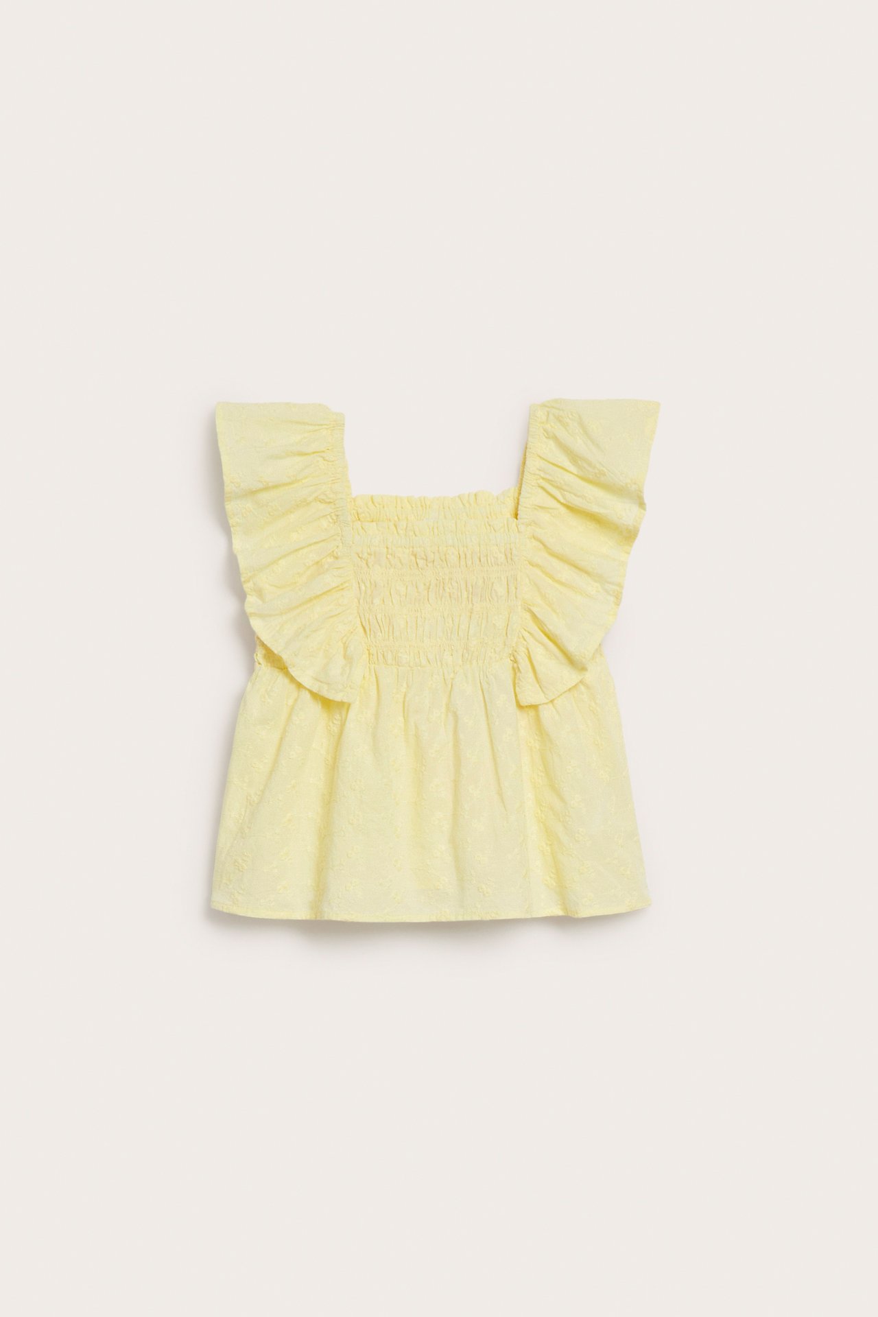 Frontvy av en barnblus i ljusgul bomull med broderat mönster, fyrkantig hals och volangärmar.