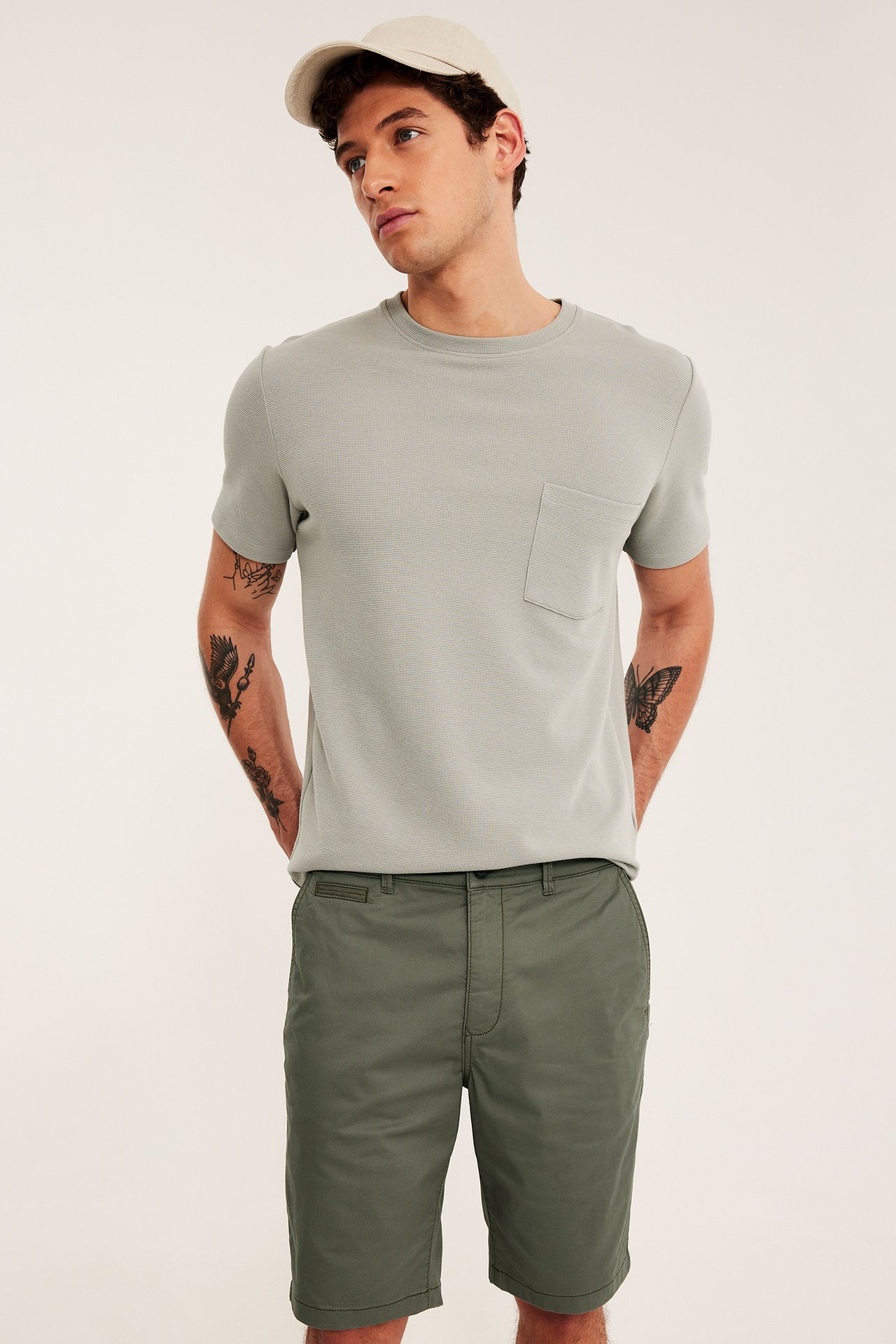 Frontvy av en man i gröna chinosshorts, en ljusgrå t-shirt med ficka och en beige keps.