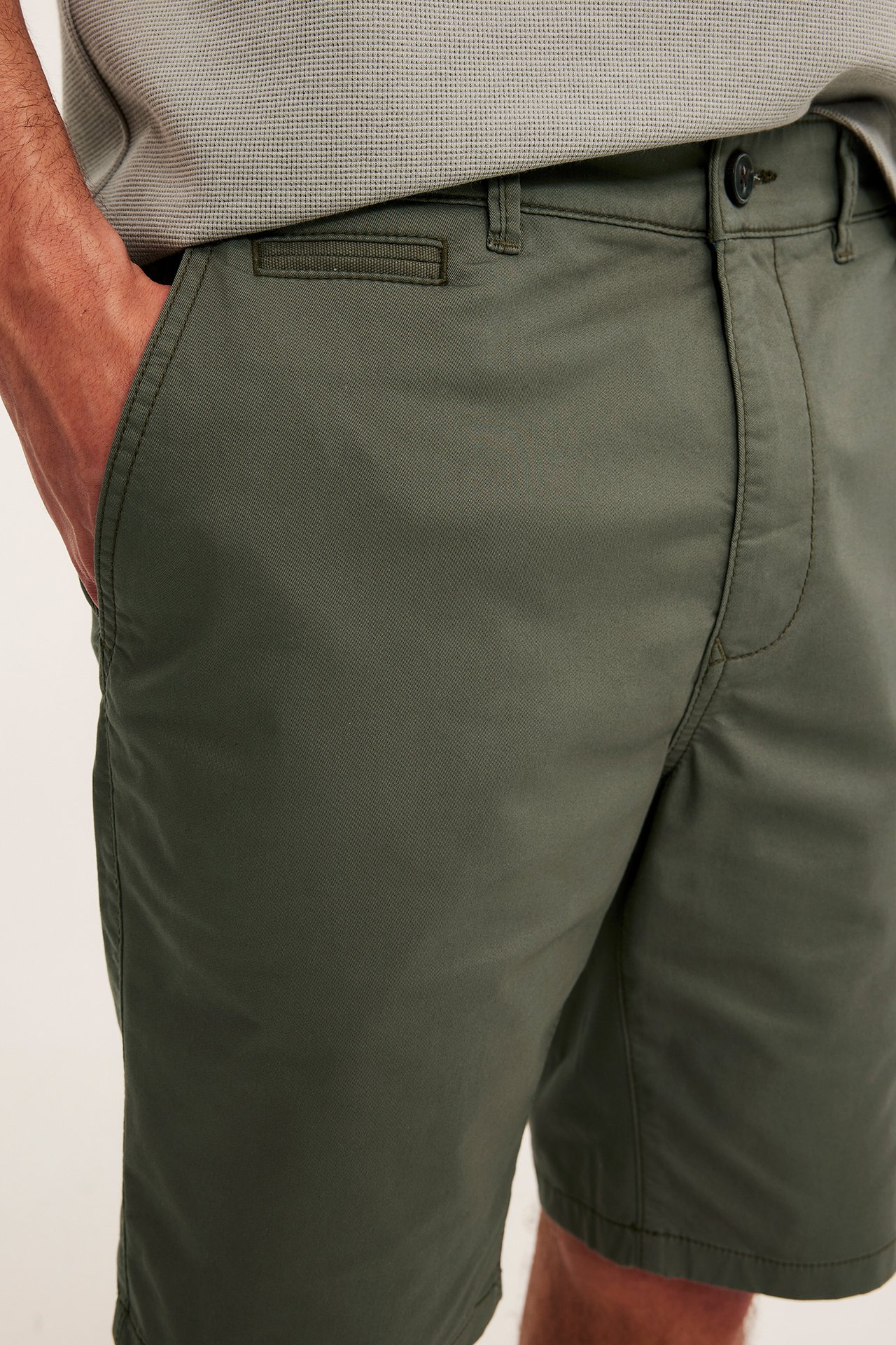 Nærbilde forfra: mann i grønne bomullschinoshorts, hånd i lommen, lysegrå pikétrøye.