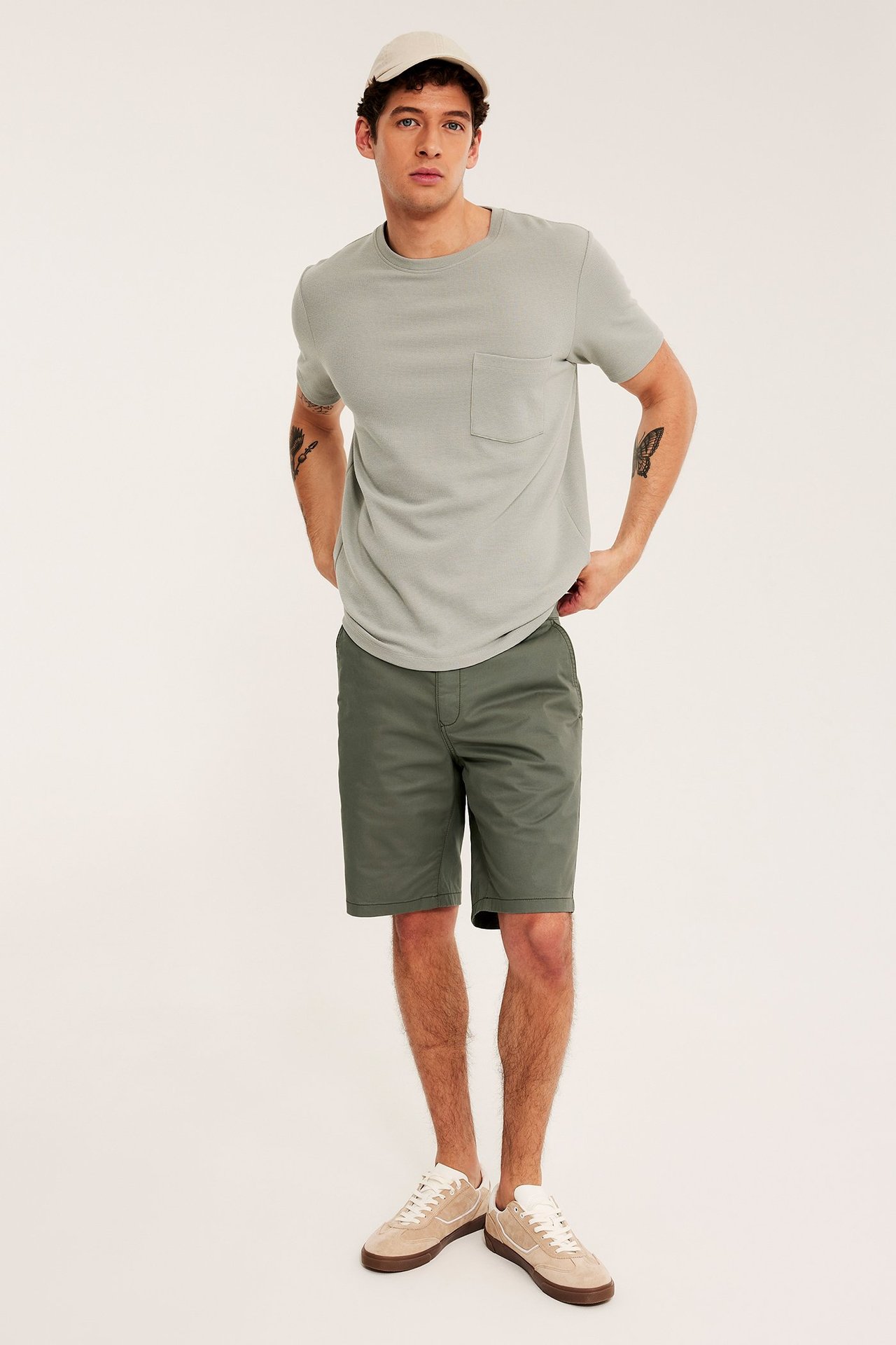 Frontvy av en man i gröna bomullstwill chinosshorts, en ljusgrå t-shirt, en beige keps och beiga sneakers.