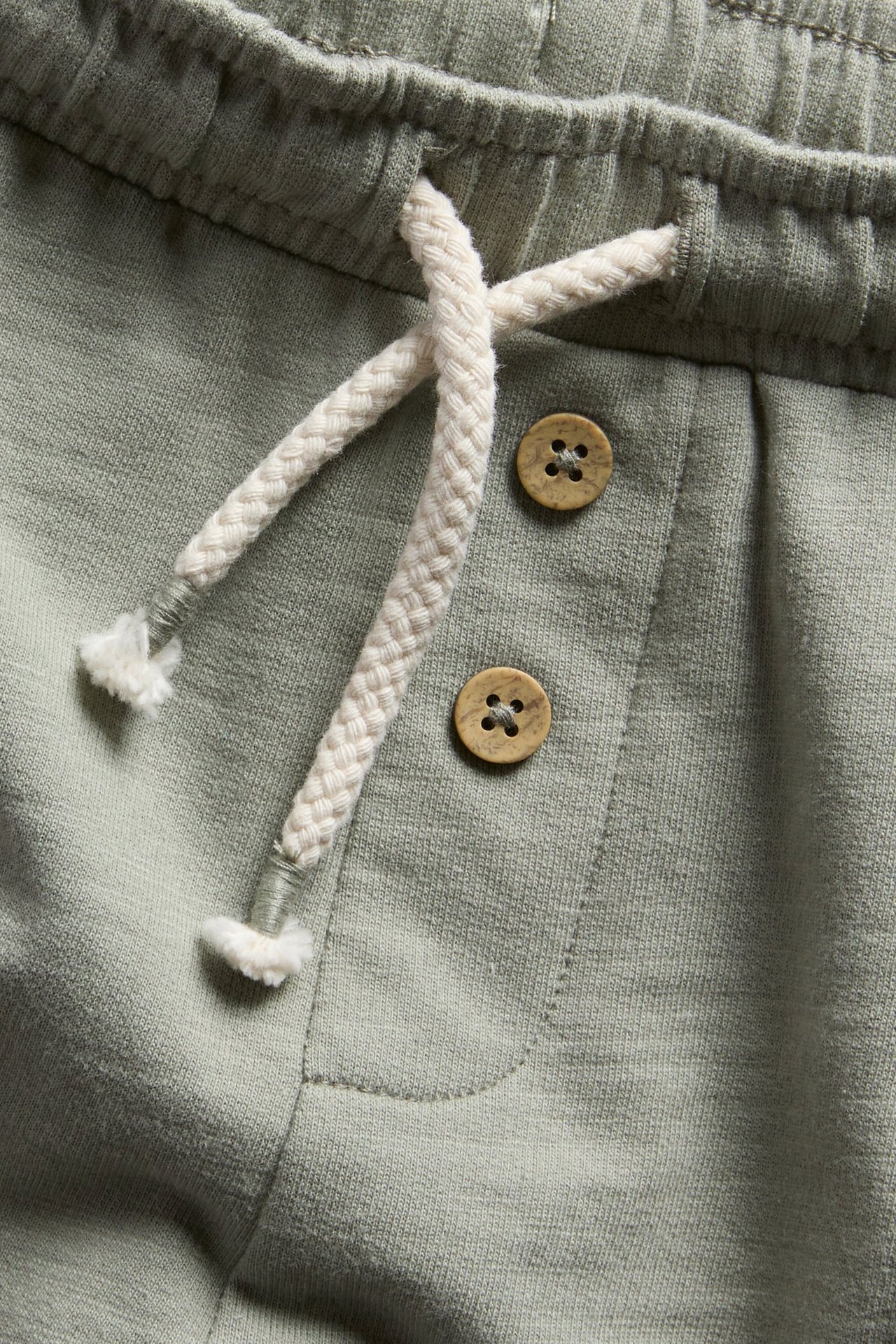 Närbild av mörkgröna shorts i slubtrikå med resårlinning, vit dragsko och två dekorativa träknappar.