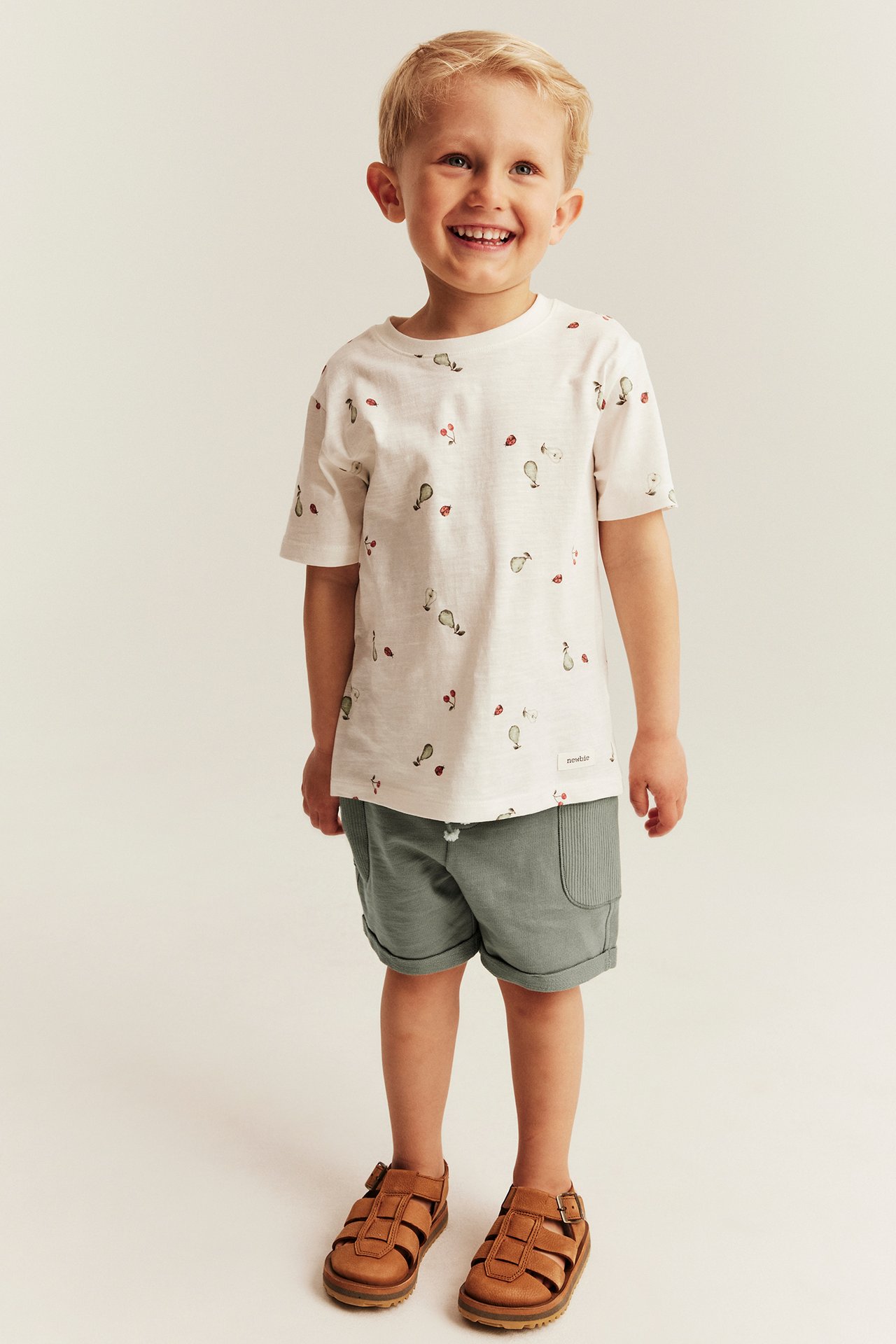 Edestä näkymä: Lapsi yllään tummanvihreät slub-trikooshortsit, valkoinen kuvioitu t-paita ja ruskeat sandaalit.