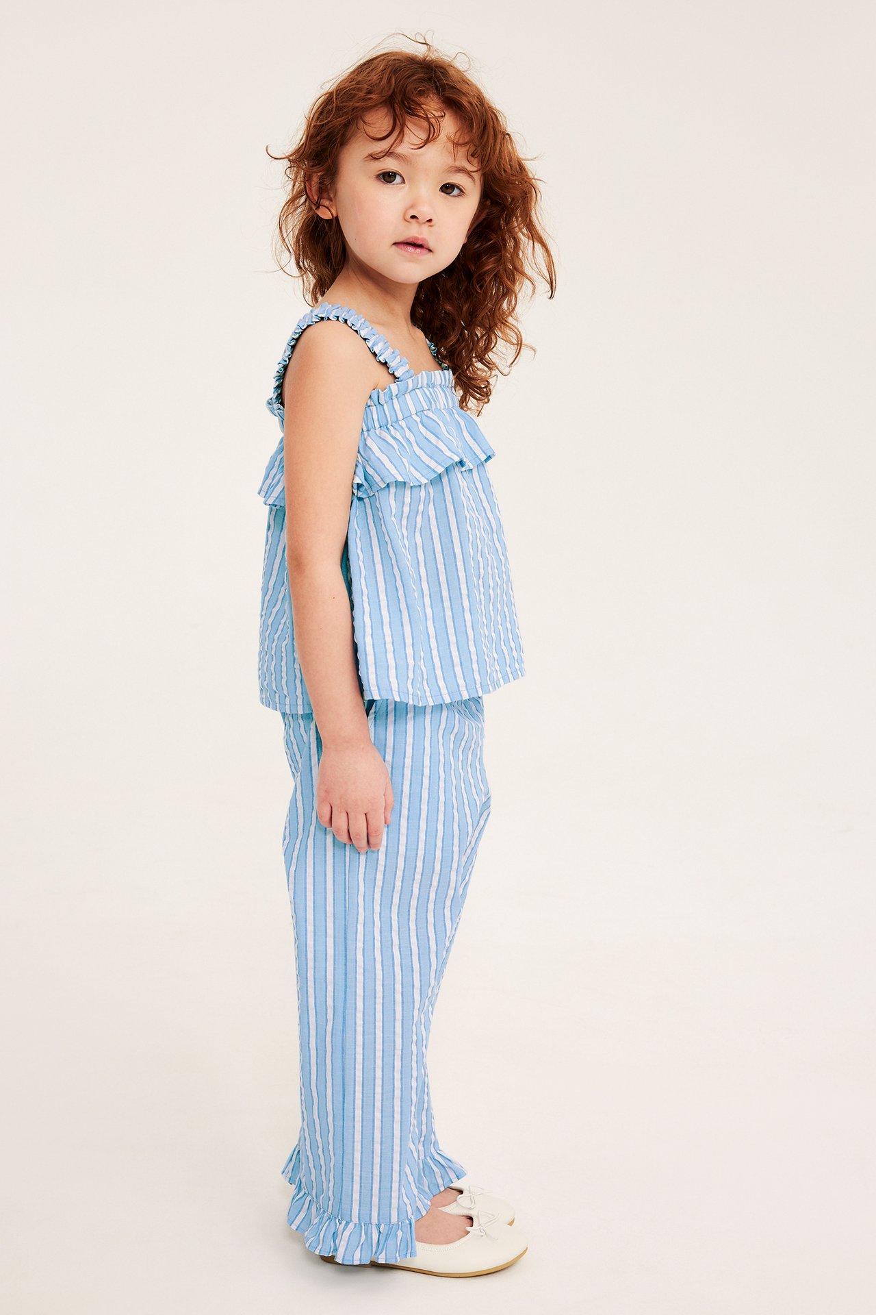 Sidovy av ett barn i blåvitrandiga bomullsbyxor, matchande volangtopp och gräddvita skor.
